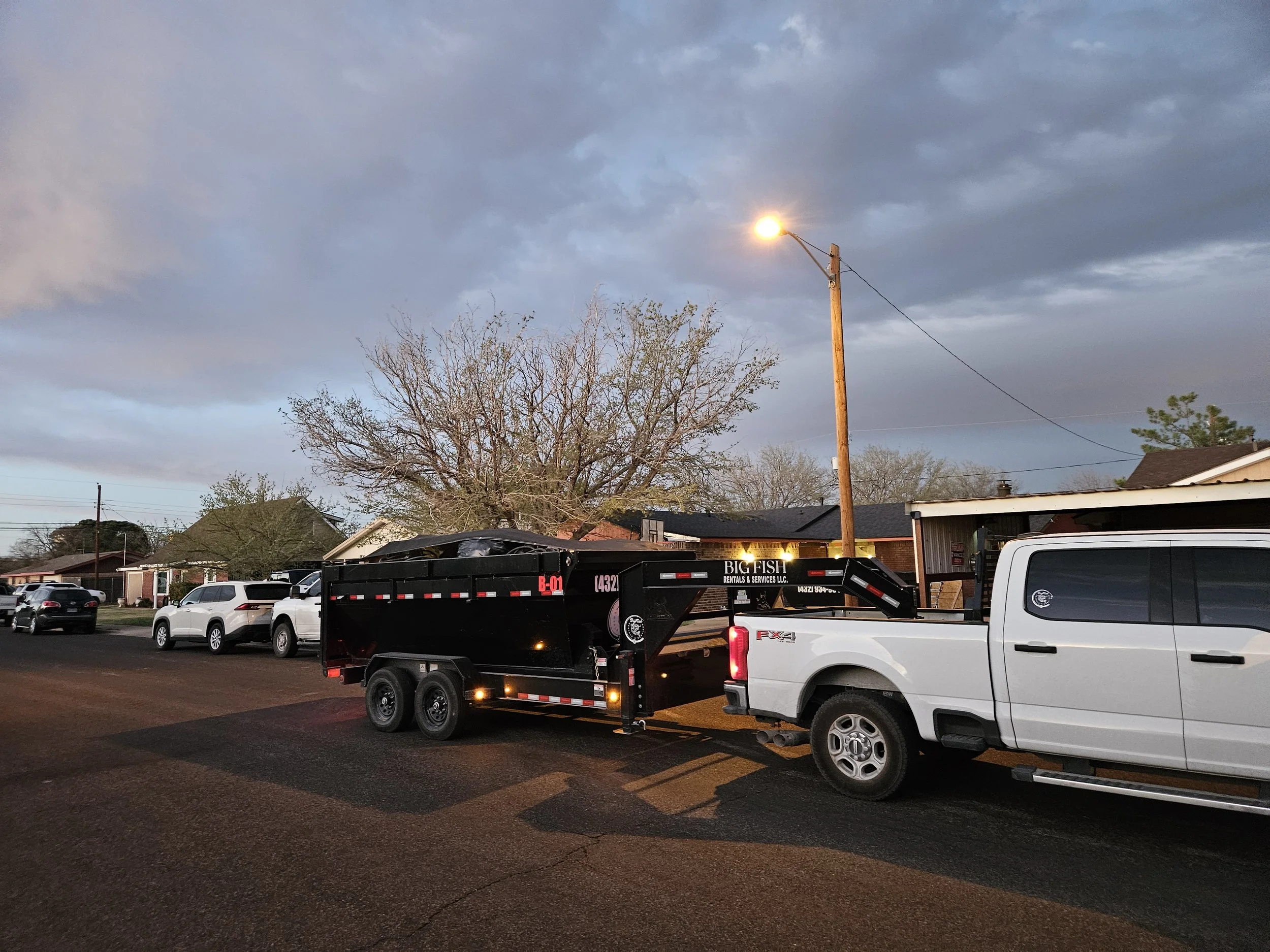 A parking lot featuring a white pickup truck and a black flatbed trailer with the words "Big Fish Rentals & Services LLC" on it. In the background, there are residential houses, leafless trees, and a streetlight turned on under a mostly cloudy sky.