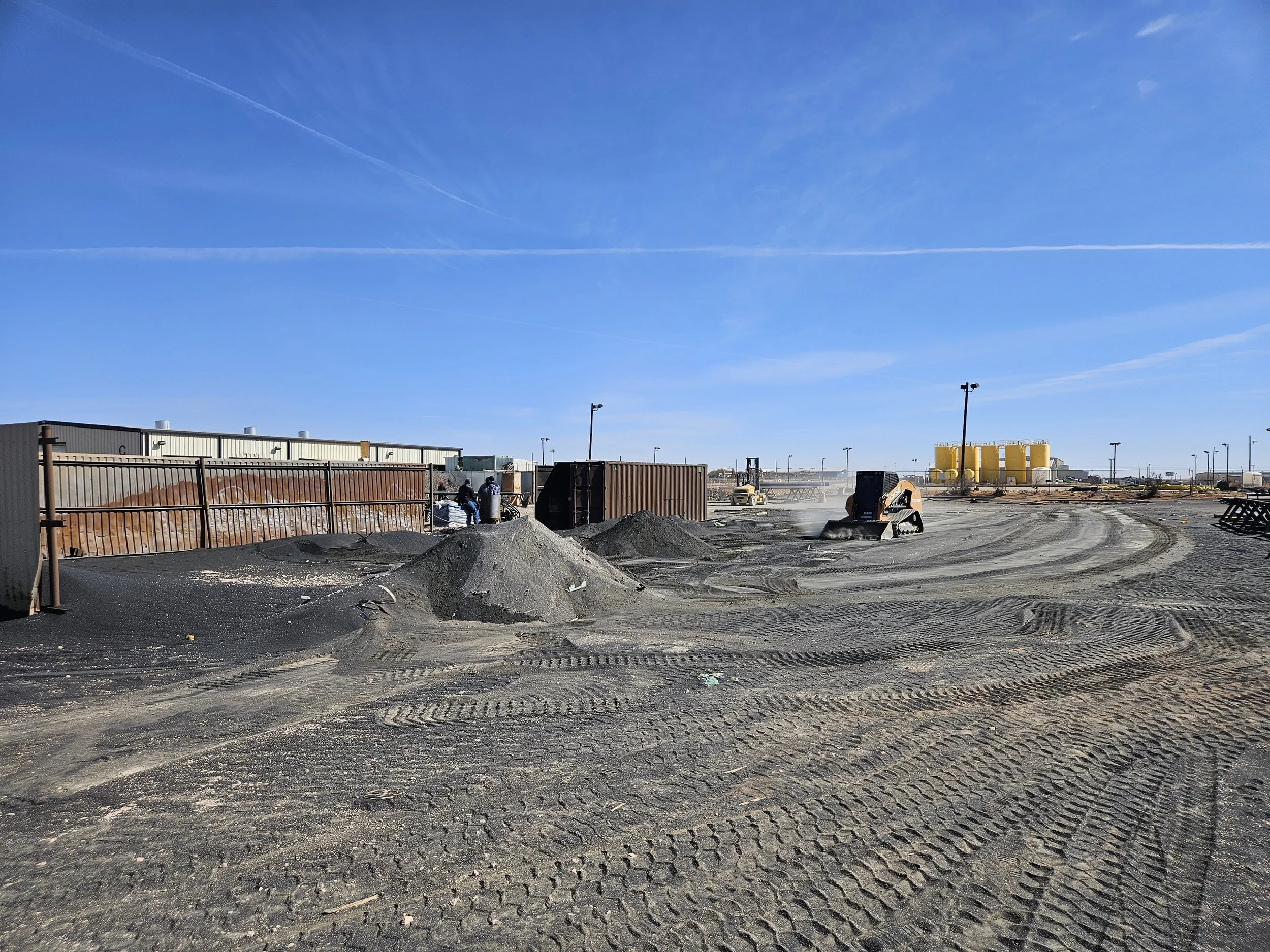 A construction site with heavy machinery, wooden fences, dirt piles, and workers under a bright blue sky with clouds and contrails.