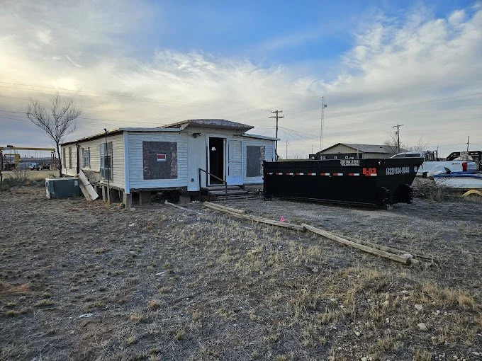 A small, weathered light blue house with boarded-up windows and a wooden ramp leading to the front door, situated in a barren, dirt yard. Next to it, there's a large black dumpster with red and white markings. Some cars are parked in the background u