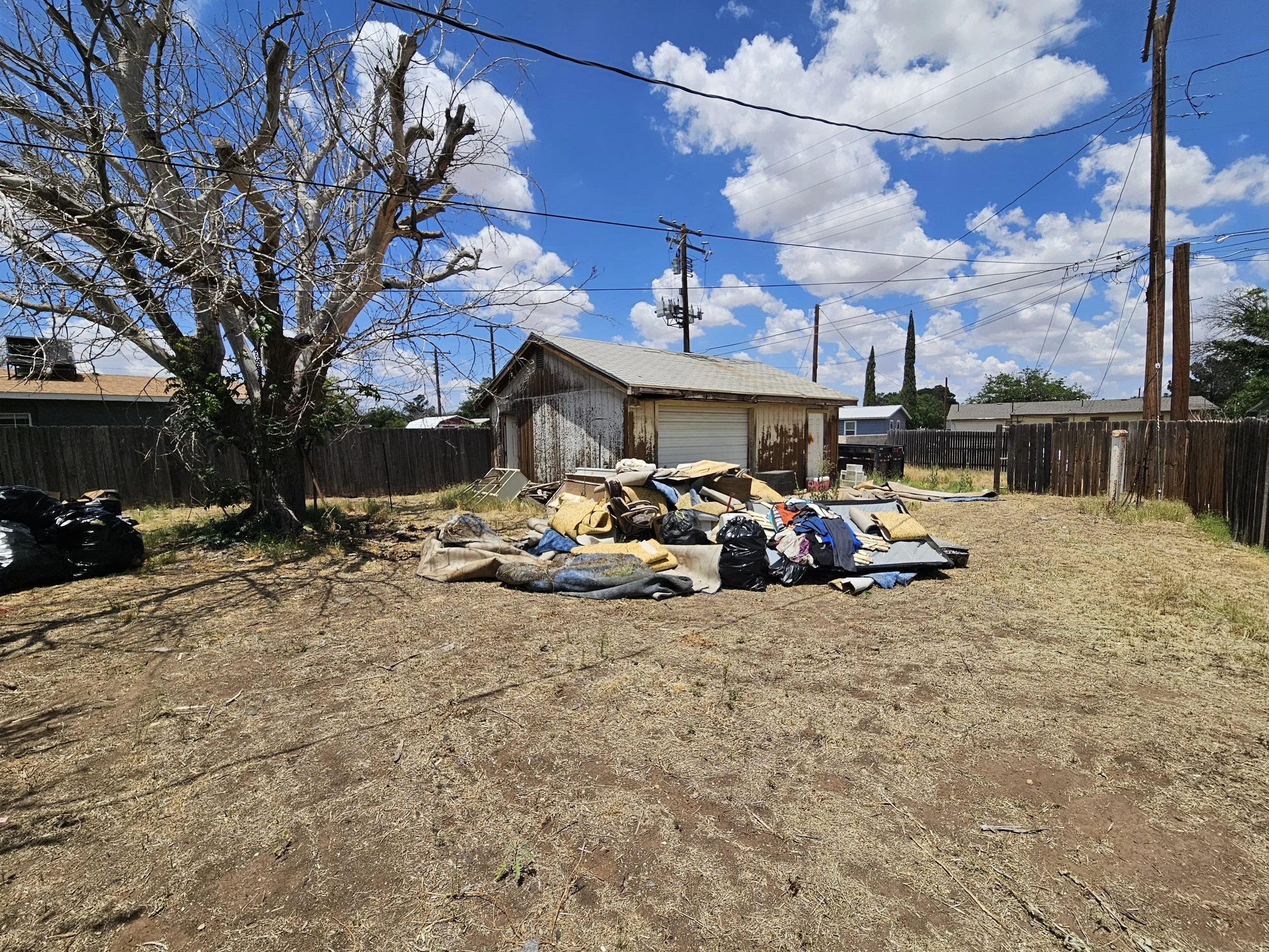 A backyard scene with a large, leafless tree, a weathered shed with a closed garage door, scattered outdoor furniture, and various items on the ground. Power lines and utility poles run across the sky, which is partly cloudy with white clouds and blu