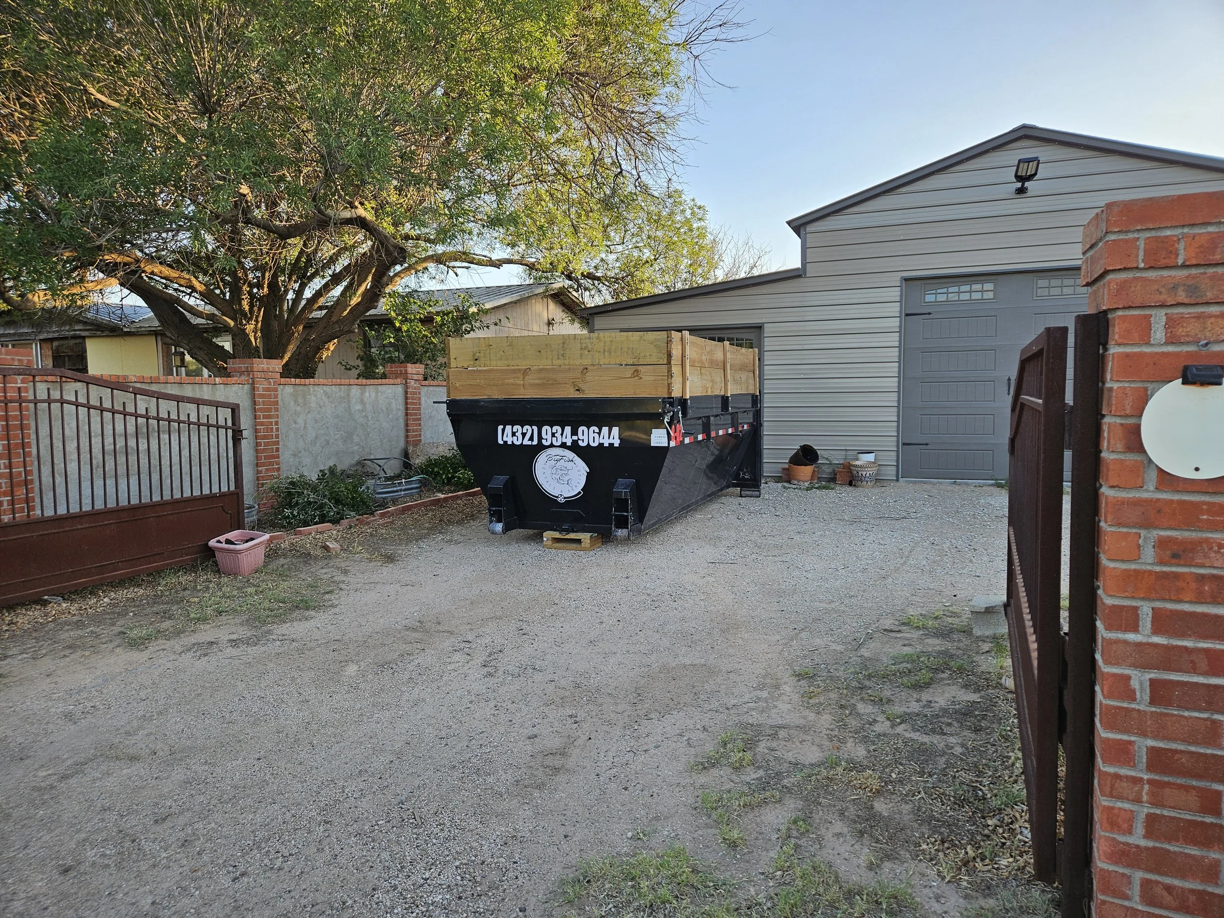 A backyard scene with a large tree, a black utility trailer with a wooden top, a gray garage with a door, and a brick and metal fence. There are some gardening pots and tools, and a gravel ground.