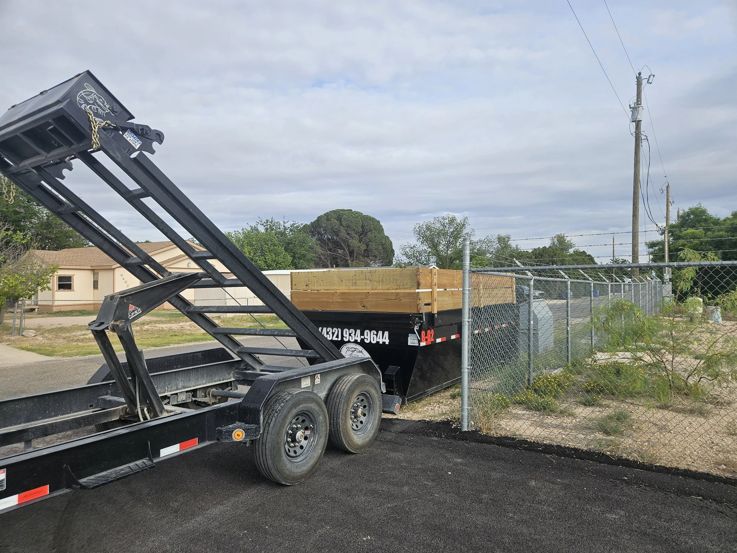 A black trailer with a wooden side panel and a logo on the side is parked on a paved street next to a chain-link fence. In the background, there are some houses, trees, utility poles, and wires under a cloudy sky.