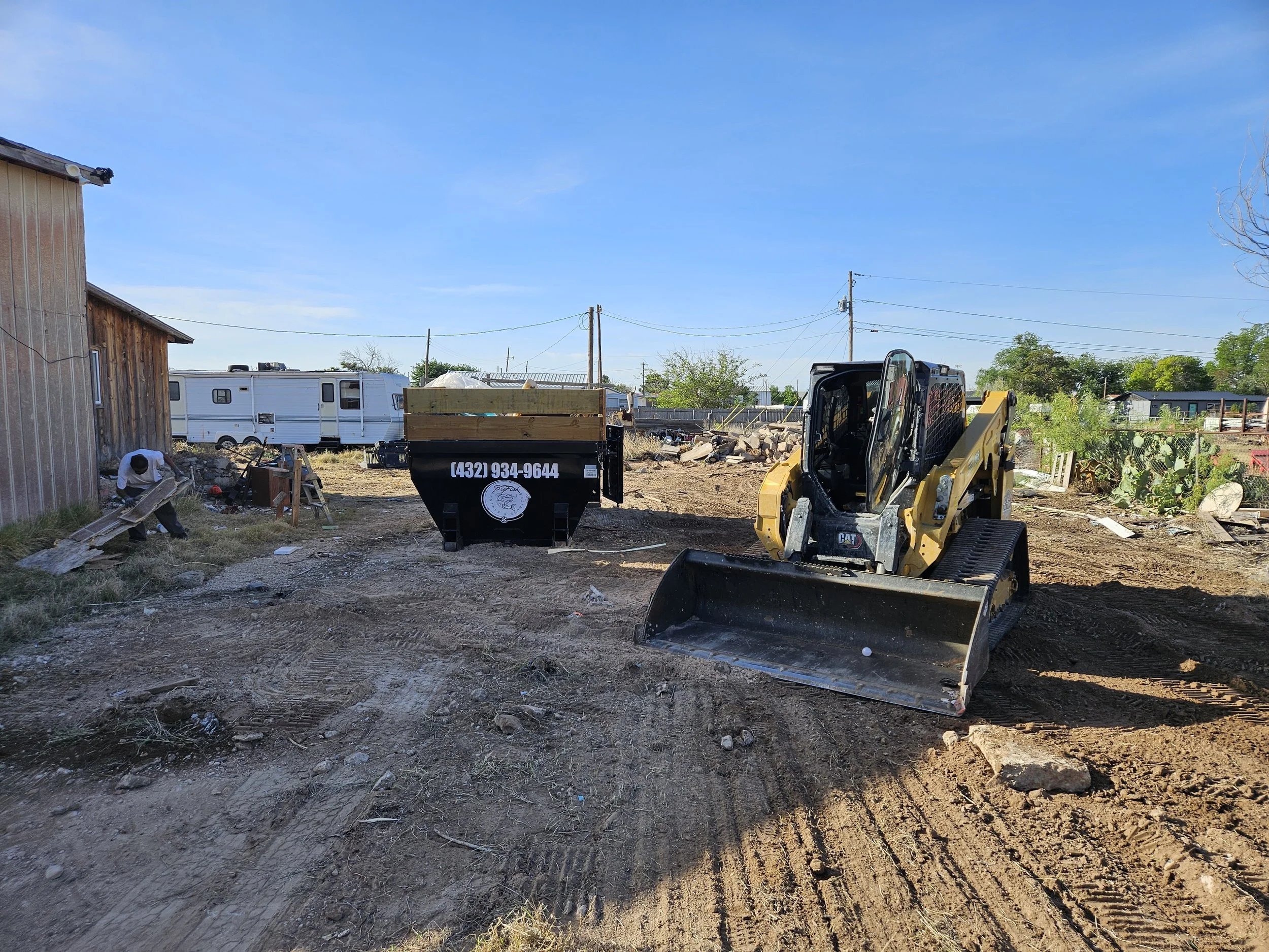 A yard under construction with a small yellow construction vehicle, a black trash bin, a white travel trailer, and scattered debris including rocks, wood, and tools. Power lines run above, and a weathered wooden building is on the left.