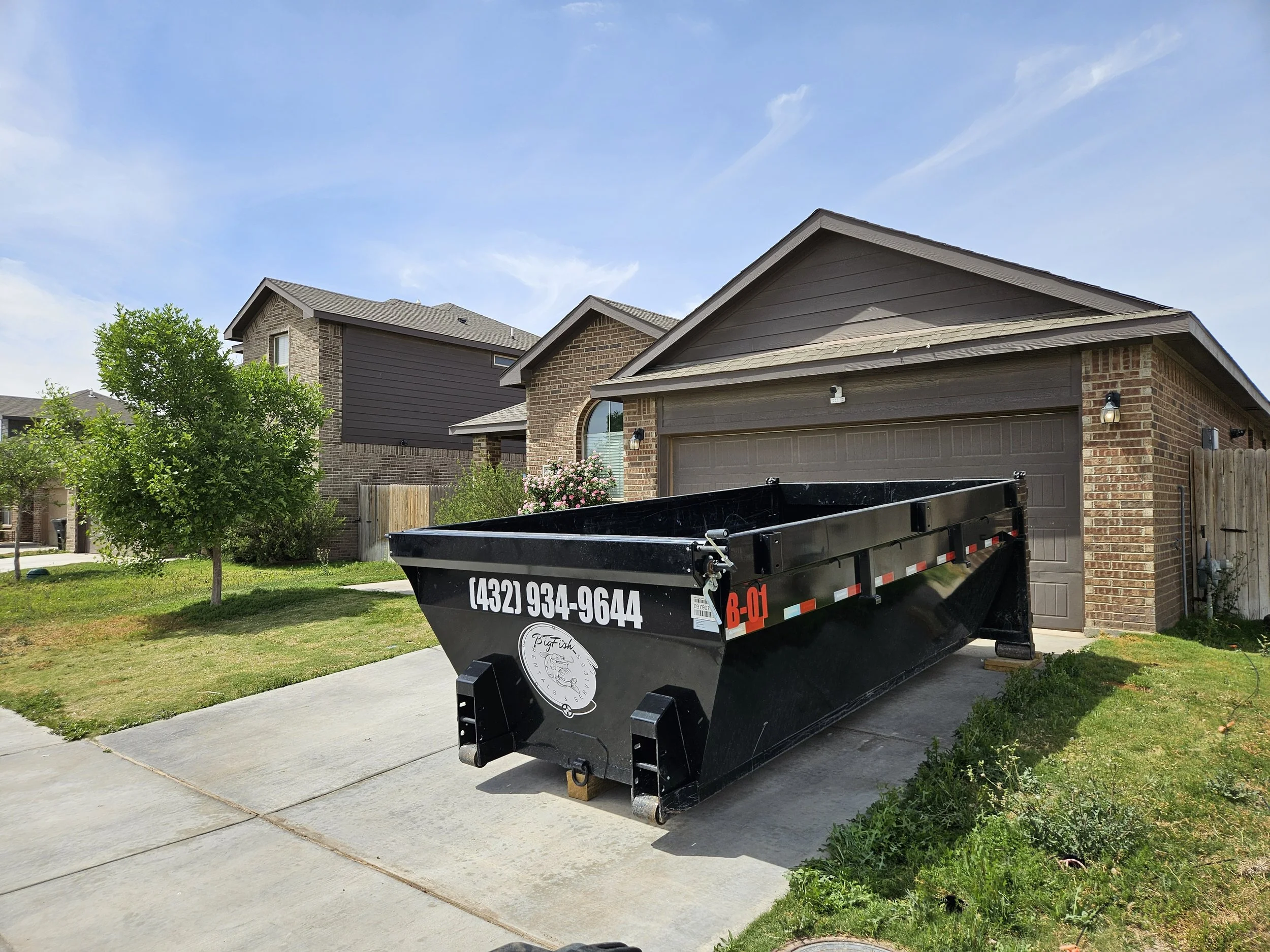 A residential neighborhood featuring brick and siding homes with well-maintained yards and trees. In the driveway, there's a black dump trailer with a phone number, logo, and some markings on it.