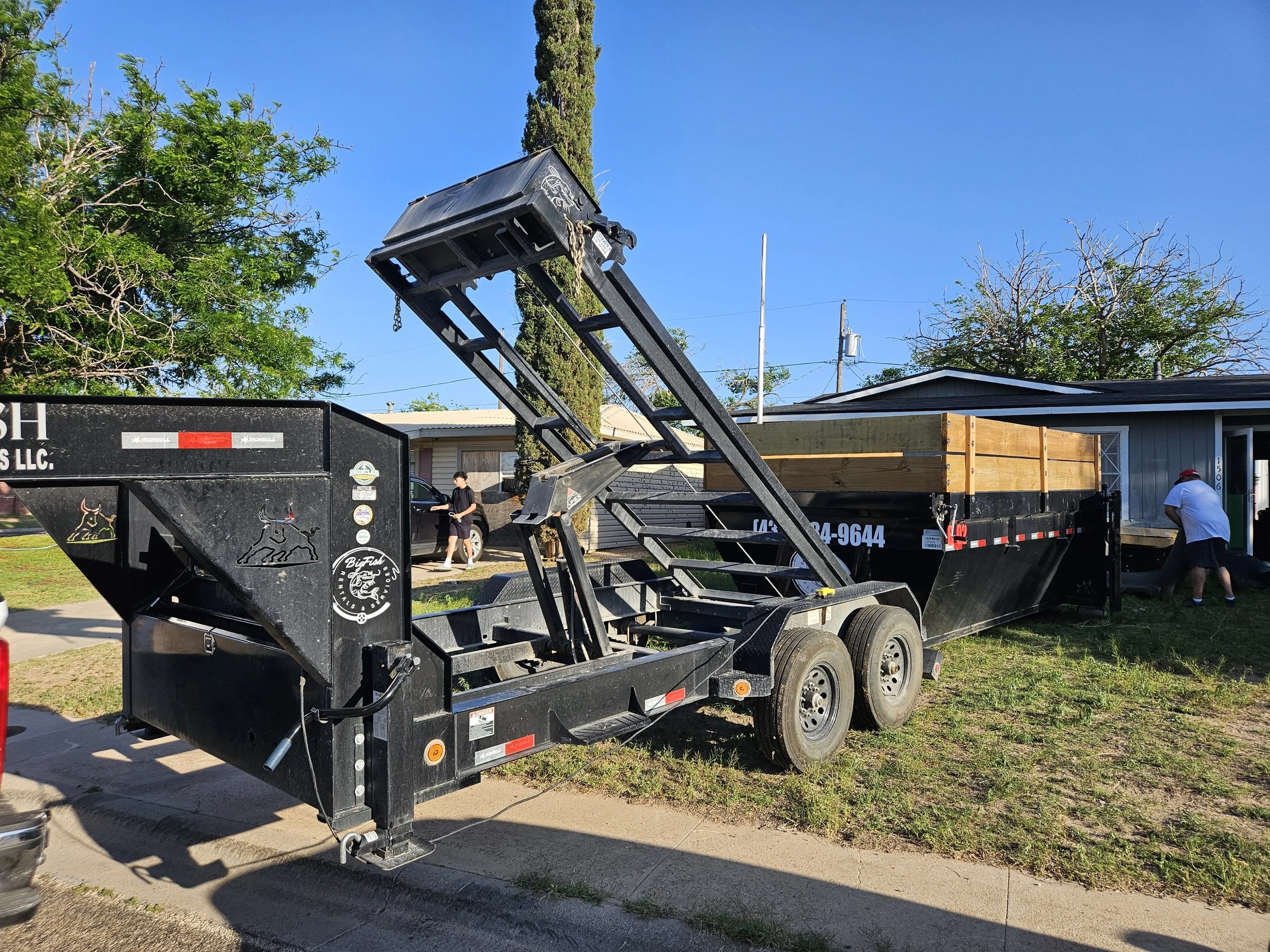 A flatbed trailer truck with a black color, parked on the side of a residential street. The trailer has a logo with a howling dog and the text "Big Fish" on the side. The truck bed has a wooden structure being loaded or unloaded with a hydraulic lift