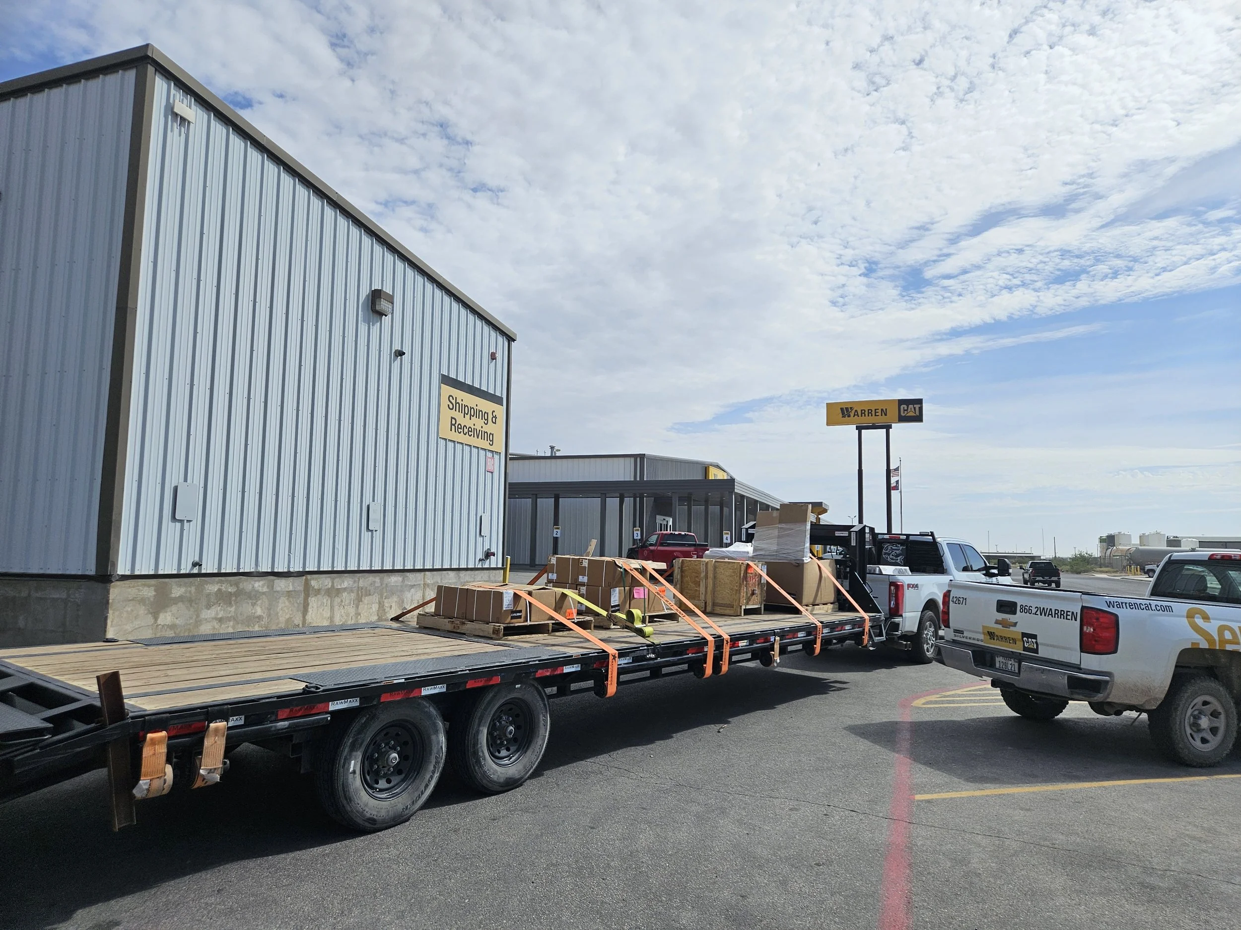 A flatbed truck carrying wooden pallets parked in a lot near a building with a sign that reads 'Shipping & Receiving.' There are several vehicles nearby, including pickup trucks with Warren CAT branding. The sky is partly cloudy.