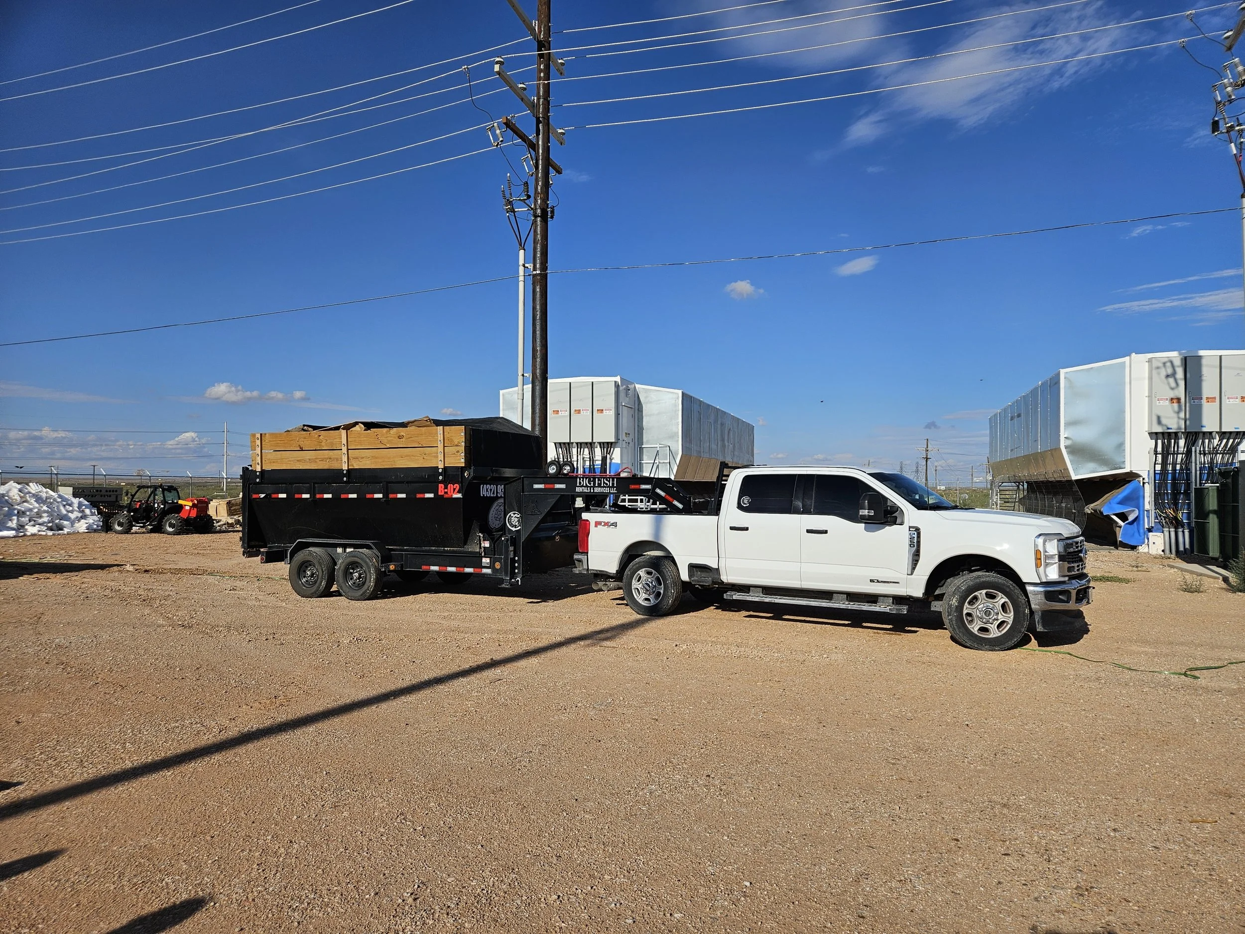 A utility pole with multiple power lines against a blue sky with some clouds, and various equipment and trucks parked on the ground below.