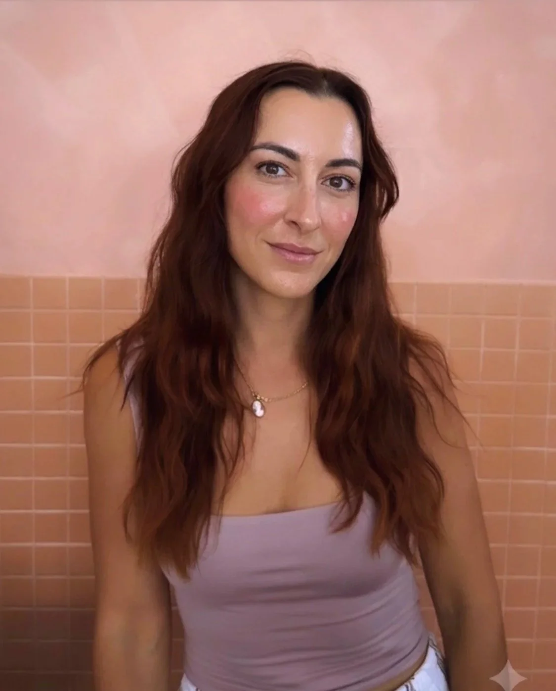 A woman with long, wavy, reddish-brown hair, wearing a light-colored tank top and a gold necklace, sitting against a peach-colored tiled wall.
