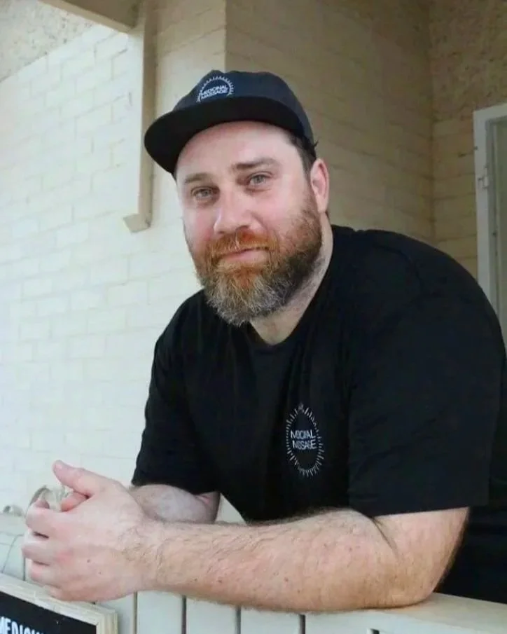 A man with a beard, wearing a black cap and black T-shirt, sitting at a table with a brick wall in the background.