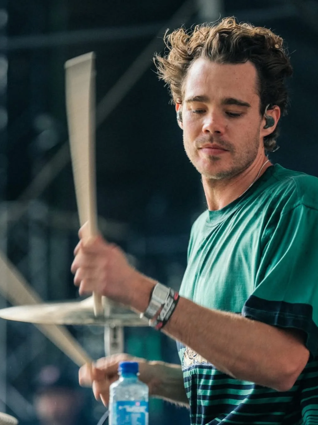A young man with curly brown hair playing drums, wearing a green shirt, holding drumsticks and wearing a wristband, with a water bottle visible in the foreground.