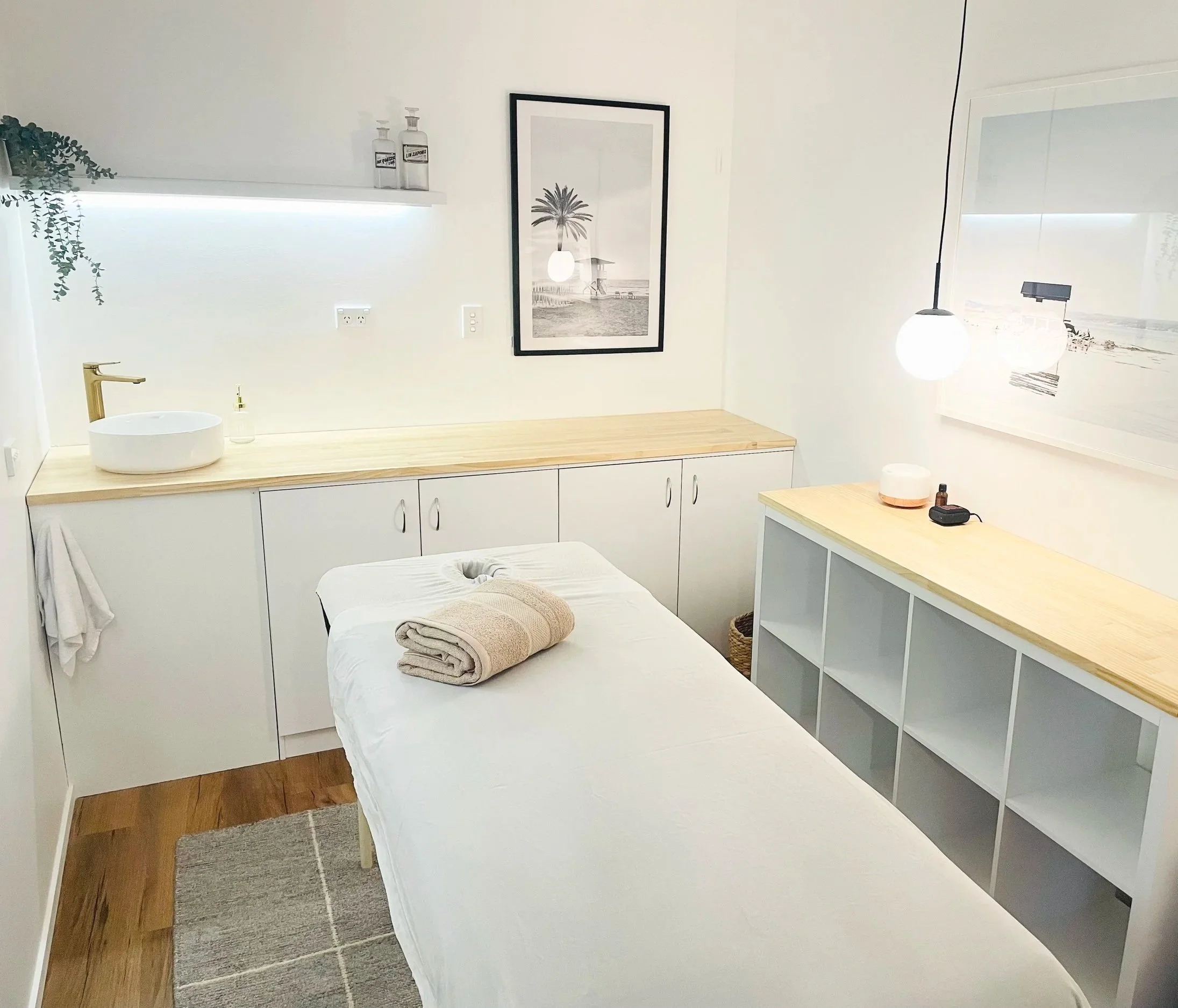 A treatment room with white walls, a massage table with beige towels, a wooden side table, a built-in white cabinet, framed beach scene art, hanging globe pendant lights, and decorative bottles and a plant on a shelf.