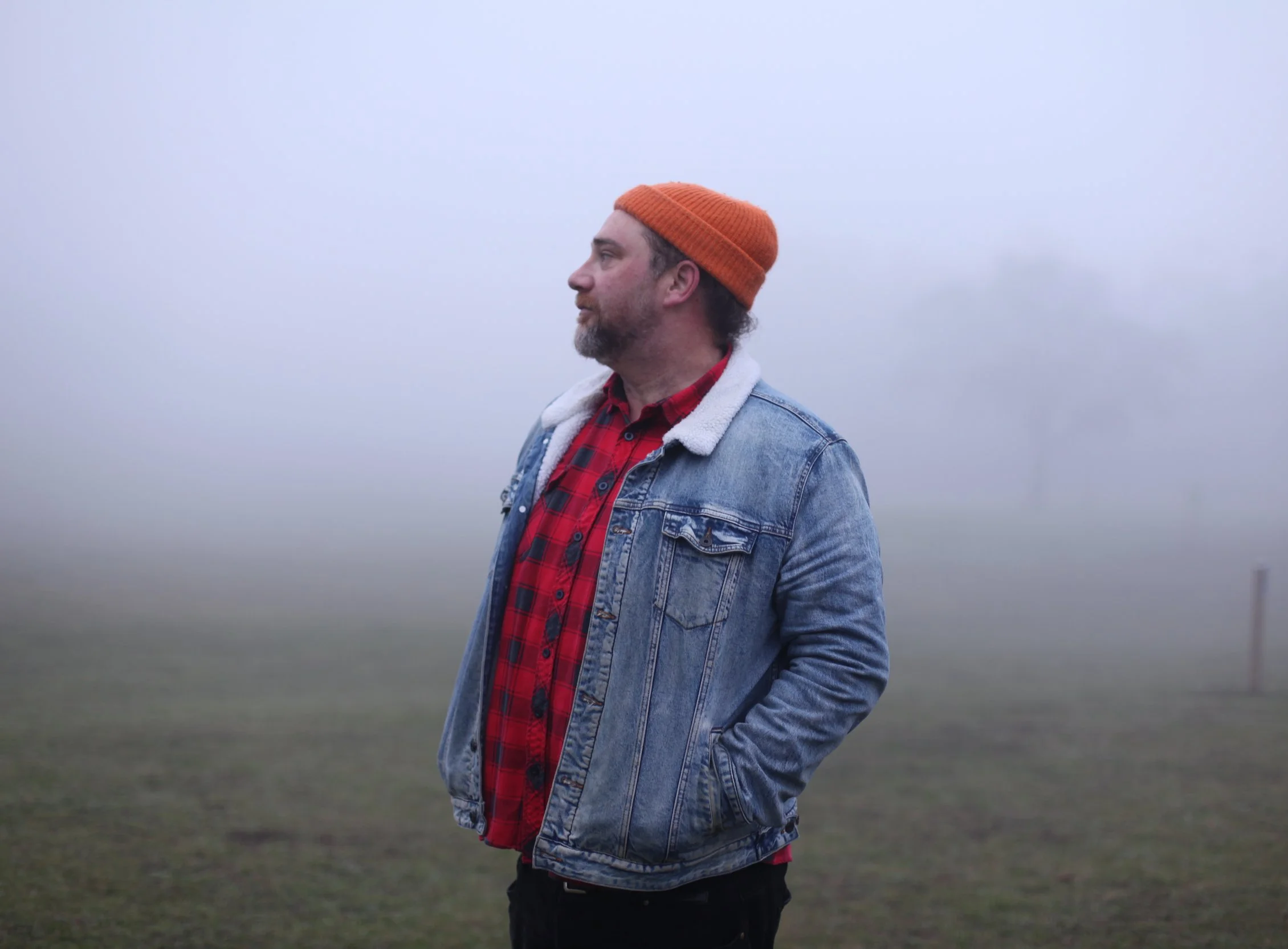 A man with a beard and mustache standing outdoors on an overcast day, wearing an orange beanie, denim jacket, red flannel shirt, and black pants, with his hands in his pockets, looking to the left.