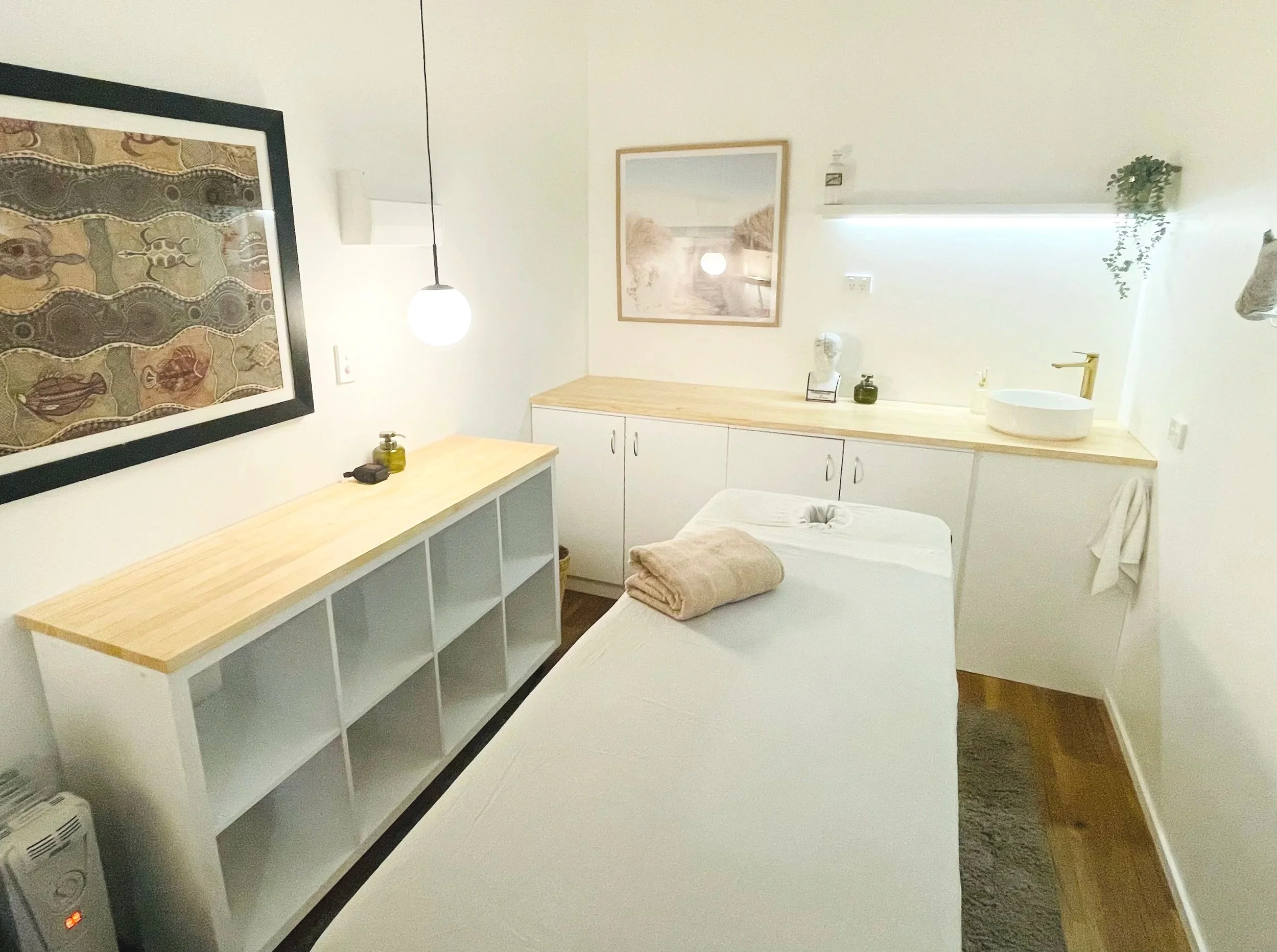 A treatment room with white walls, a massage table with beige towels, a wooden side table, a built-in white cabinet, framed beach scene art, hanging globe pendant lights, and decorative bottles and a plant on a shelf.