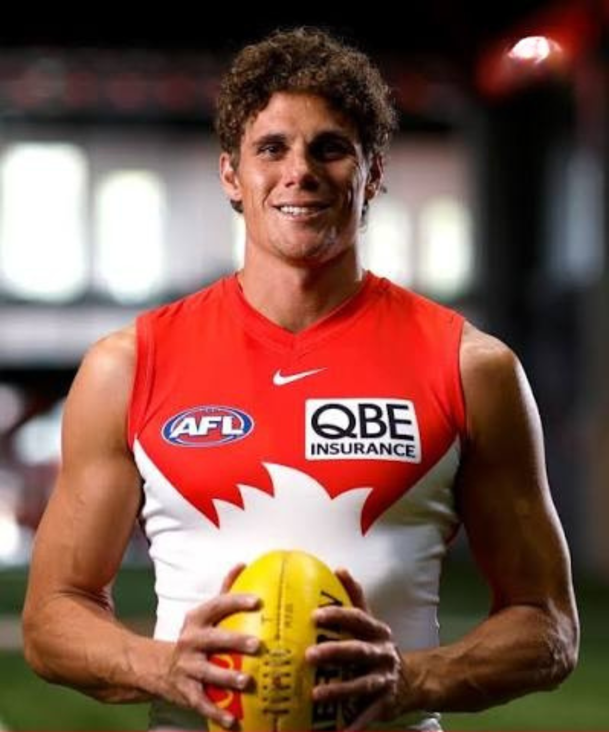 A male athlete wearing a red sports jersey with Australian Football League (AFL) and QBE Insurance logos, holding a yellow football, smiling, in an indoor sports arena.