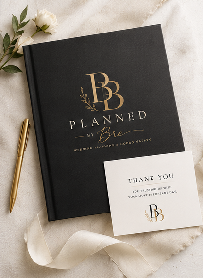 Black wedding planning book with gold lettering, a white thank you card, white flowers, and a gold pen on a beige fabric surface.