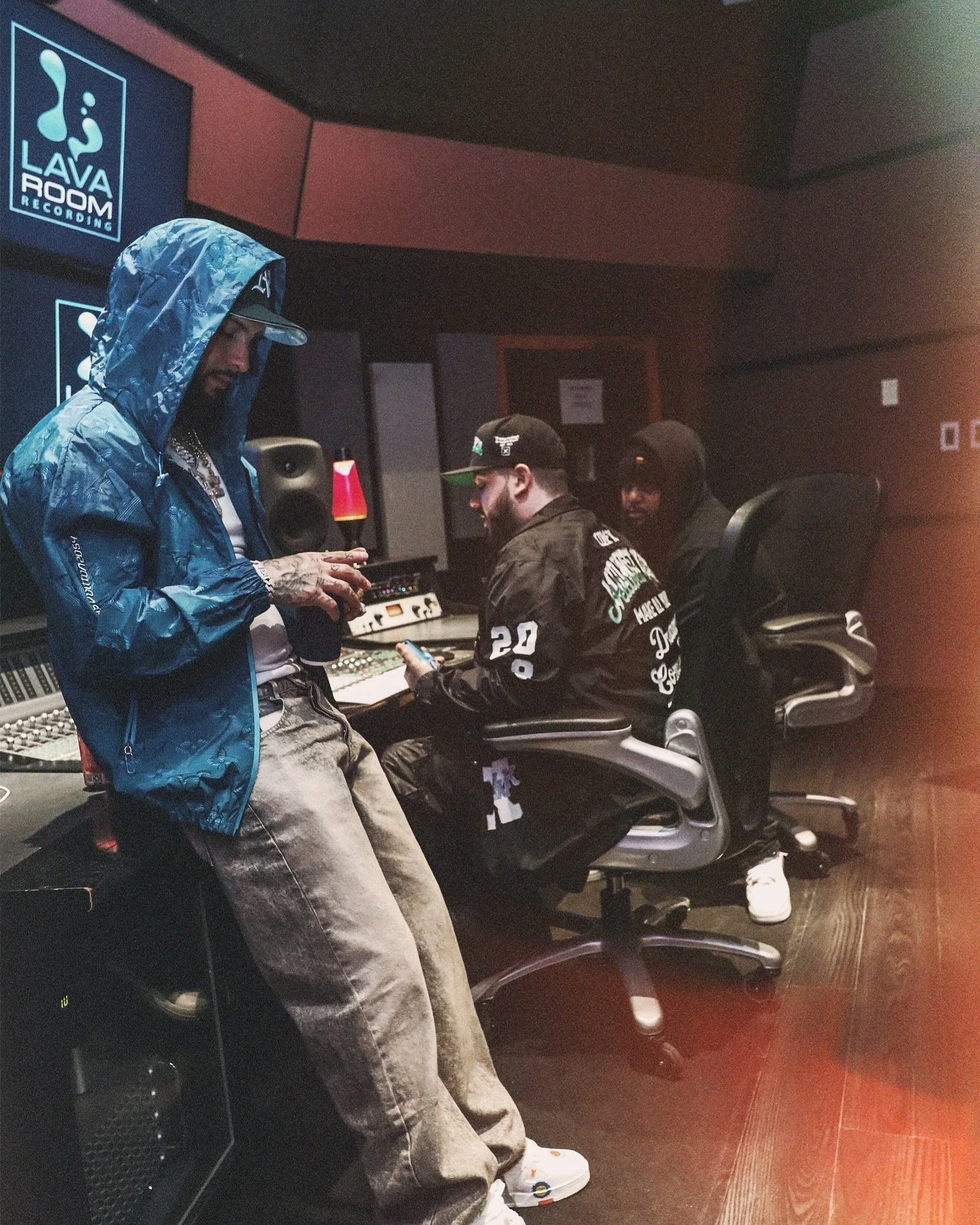 In a recording studio, three men are working at audio equipment. One man, wearing a blue patterned hooded jacket and beige pants, stands and is looking at his phone. Seated are two other men, one wearing a black cap and jacket with white text, and the other in a black hoodie and glasses. Behind them are large speakers, computer monitors, and studio equipment.