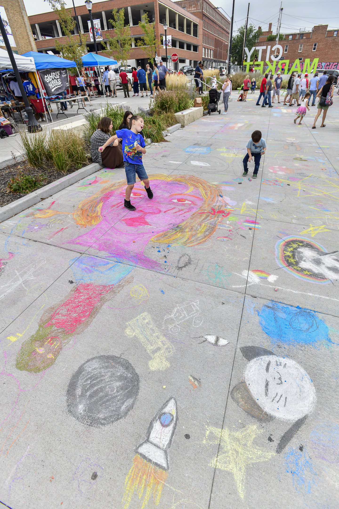 Children creating colorful chalk art on city sidewalk, with onlookers and vendors on the street behind.