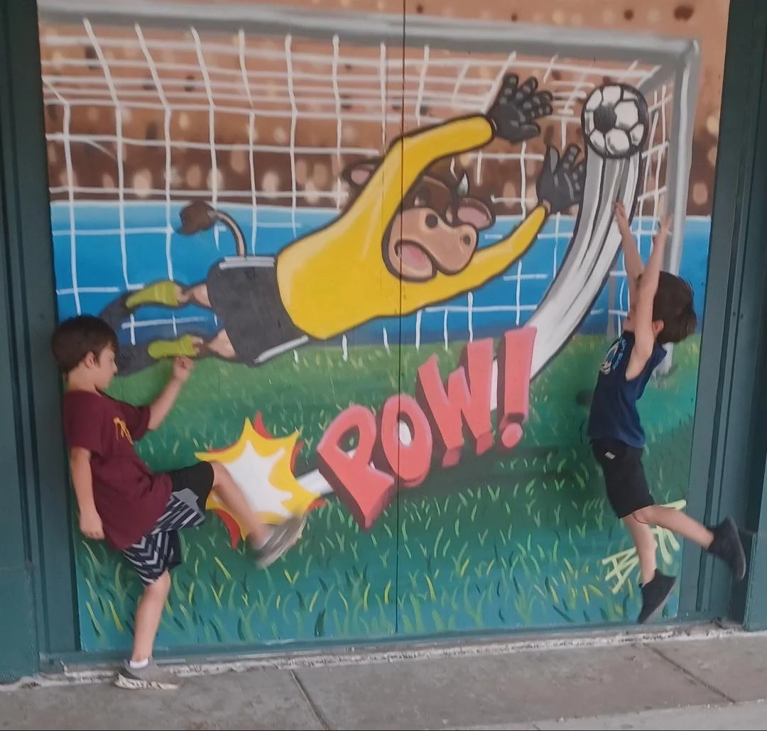Two children play in front of a colorful soccer goal mural, with a cartoon goalie diving for a ball labeled 'POW!', and one child is kicking a small ball while the other reaches up.
