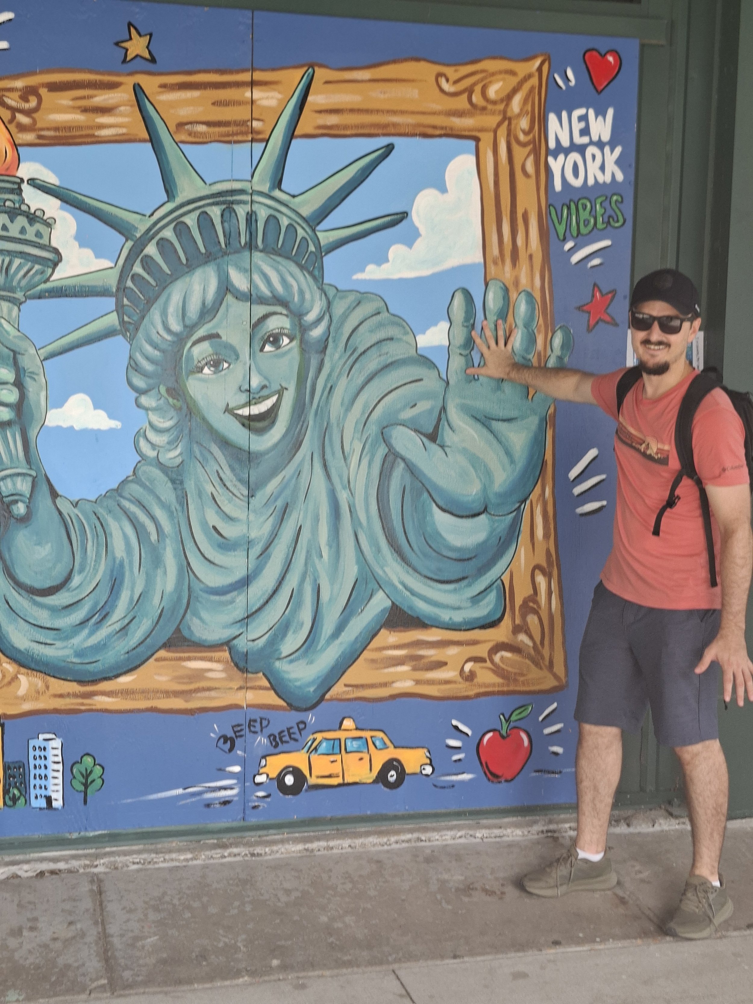 Colorful mural featuring the Statue of Liberty, a yellow taxi, a red apple, and city buildings with a blue sky and clouds in the background. A man with sunglasses, a red t-shirt, black shorts, and a backpack is touching the mural near the statue's hand.
