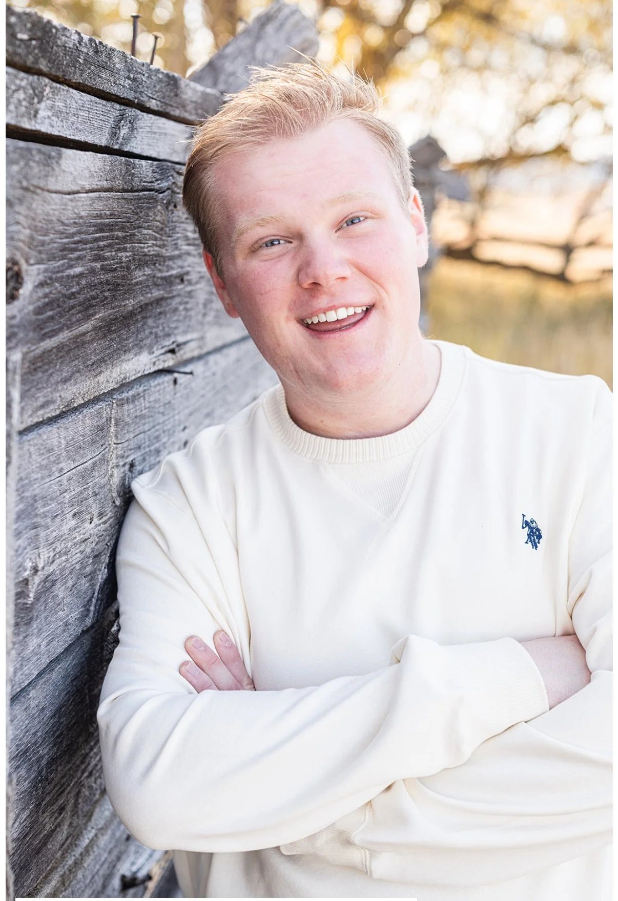 A young man with blonde hair and blue eyes smiling, leaning against a weathered wooden fence outdoors during fall, wearing a cream-colored sweatshirt with a logo on the chest.