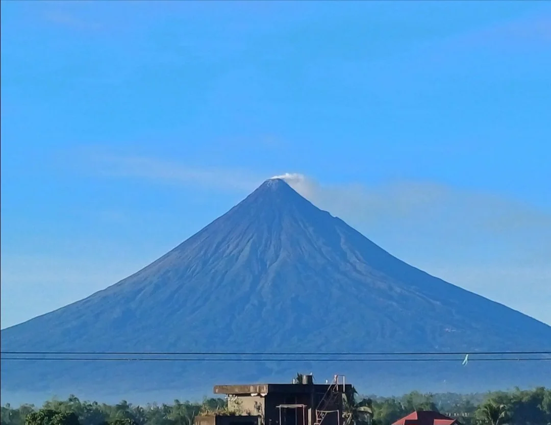 Snow-capped volcano with a small cloud at its peak, seen from a flat area with some buildings and greenery in the foreground, under a blue sky.