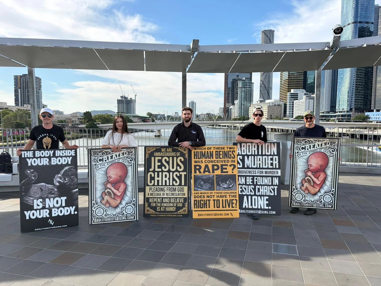 Group of five protesters standing outdoors holding signs with anti-abortion messages, with city skyscrapers and a river in the background.