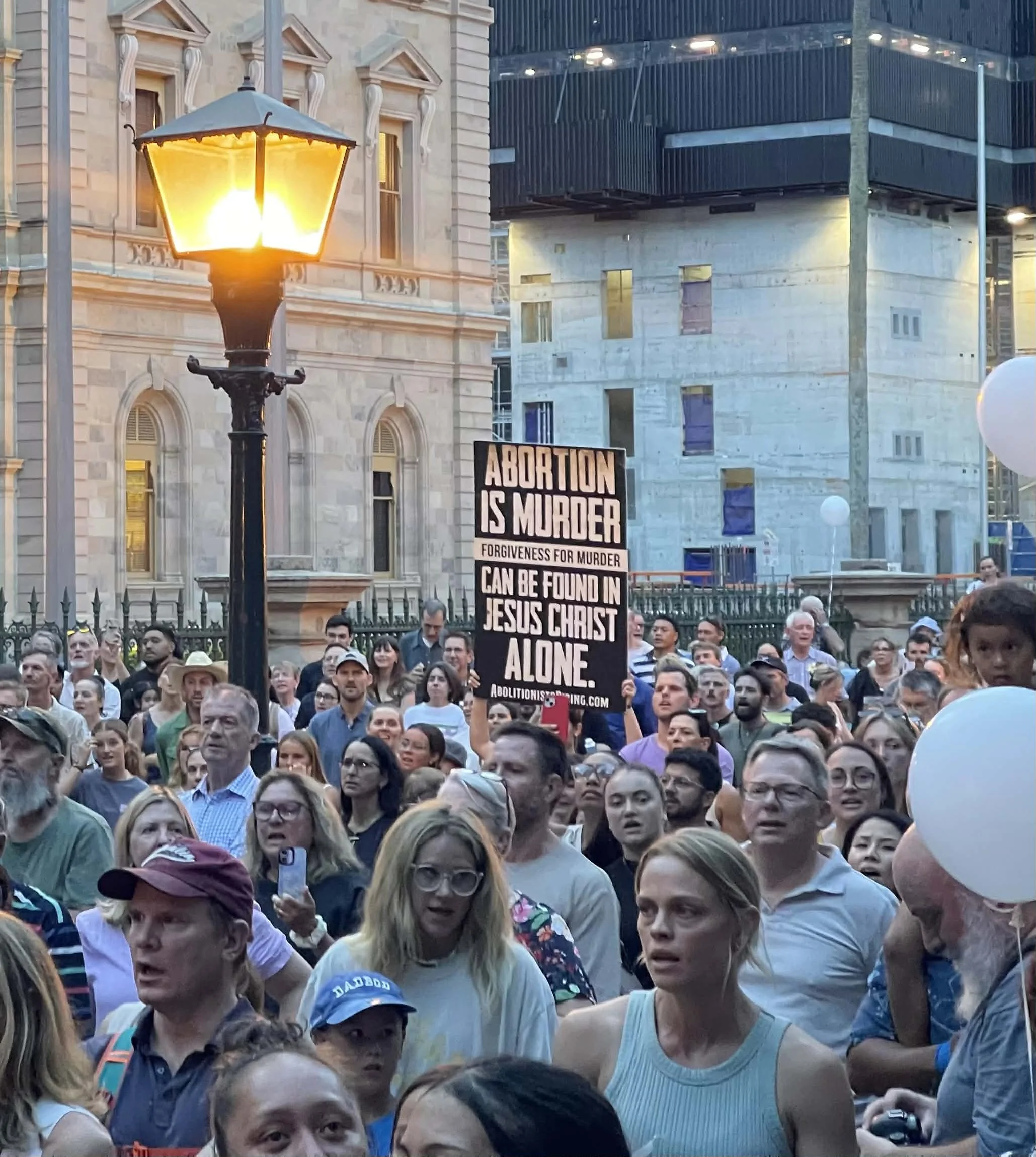A large crowd of people gathered outside in an urban area with historic and modern buildings. Some individuals hold signs, one of which reads 'ABORTION IS MURDER.' The scene appears to be a protest or rally.