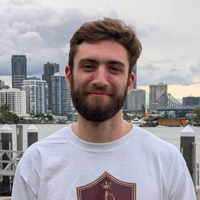 Young man with a beard smiling outdoors with city skyline and bridge in the background.