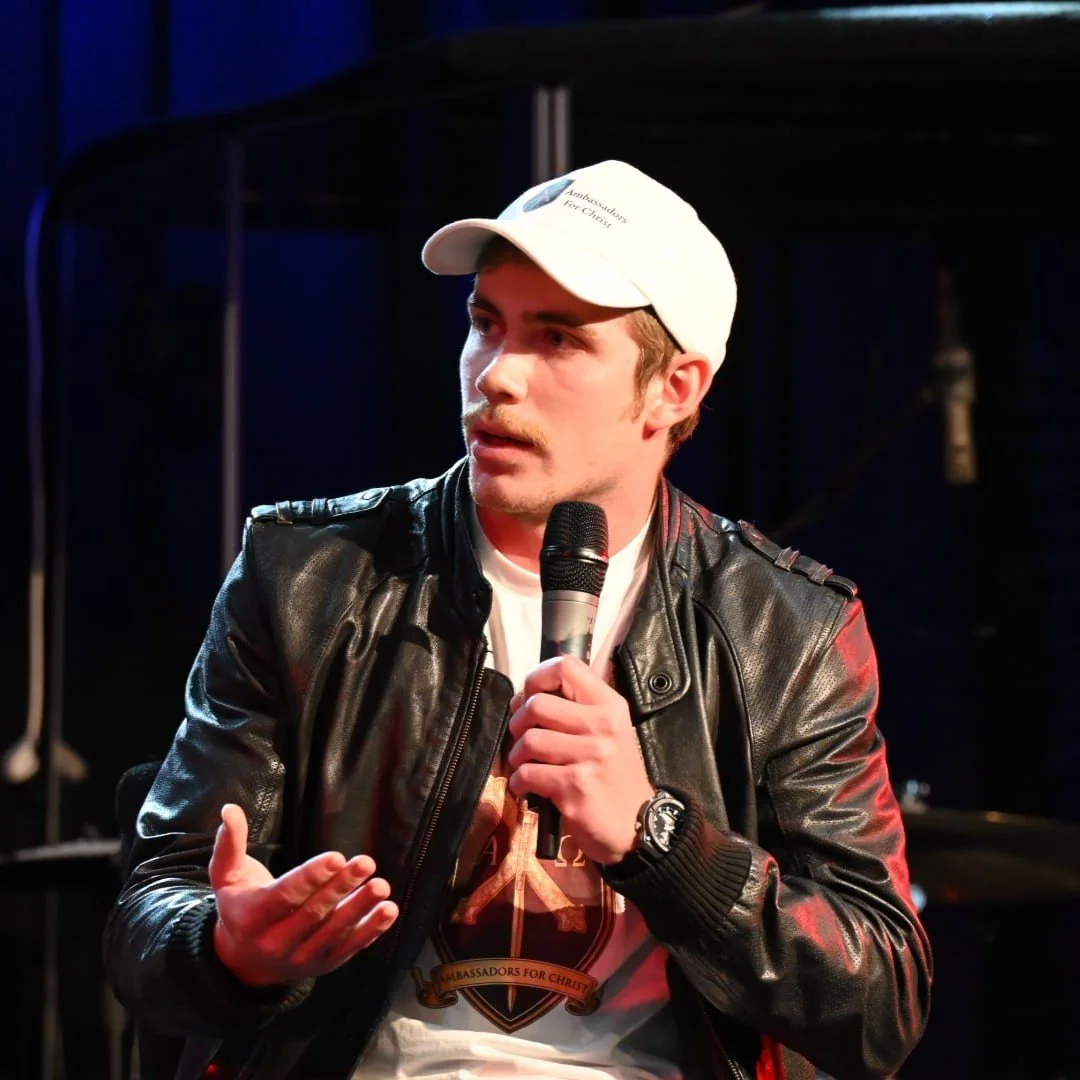 A young man in a white cap and black leather jacket holds a microphone, speaking or performing on a stage.