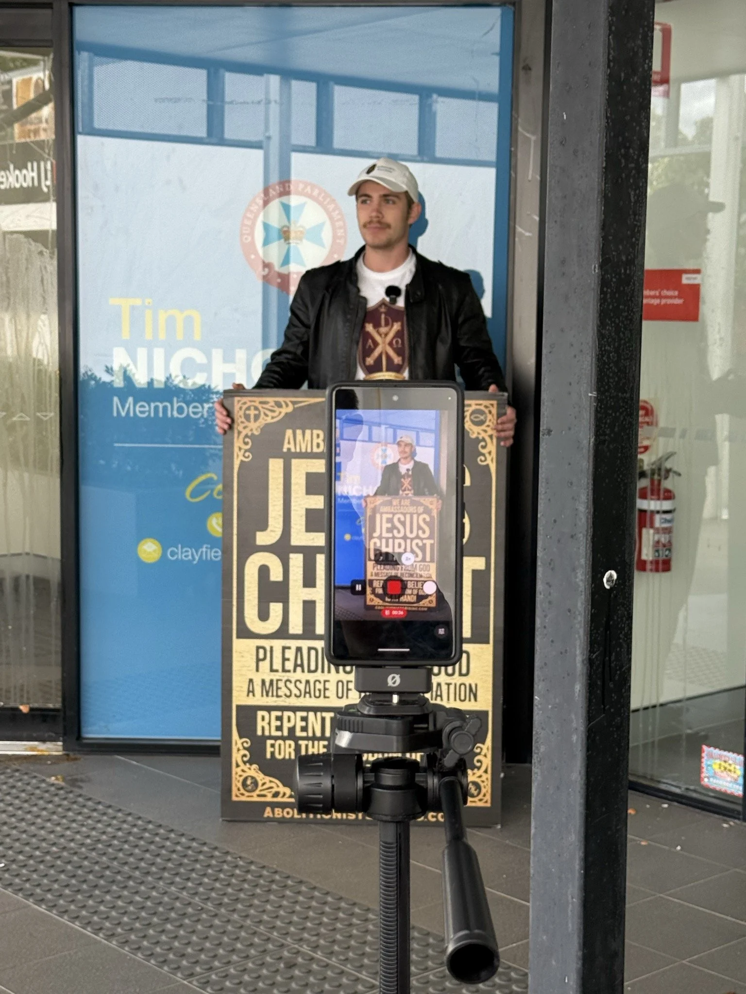 A man standing outside a building in front of a large poster displays a protest message about ending abortion and promoting religion. The man holds a cardboard sign that promotes Jesus Christ and urges repentance. There is a smartphone attached to a tripod in front of him, recording the scene.