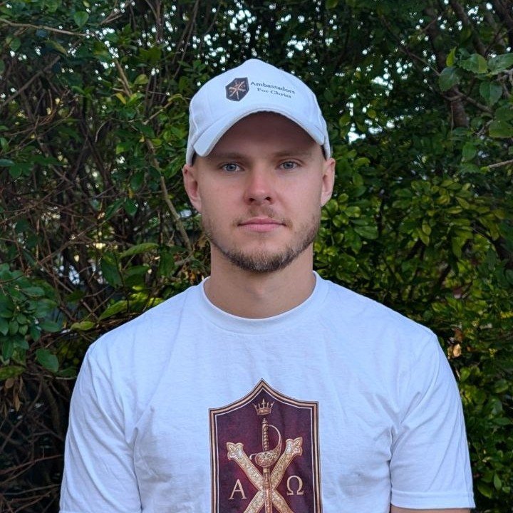 A young man with fair skin, blue eyes, and light facial hair, standing outdoors with green foliage in the background. He is wearing a light gray baseball cap with a logo and text that says "Ambassadeurs de la rédemption" and a white t-shirt with a crest featuring a serpent, a sword, and Greek letters Alpha and Omega.