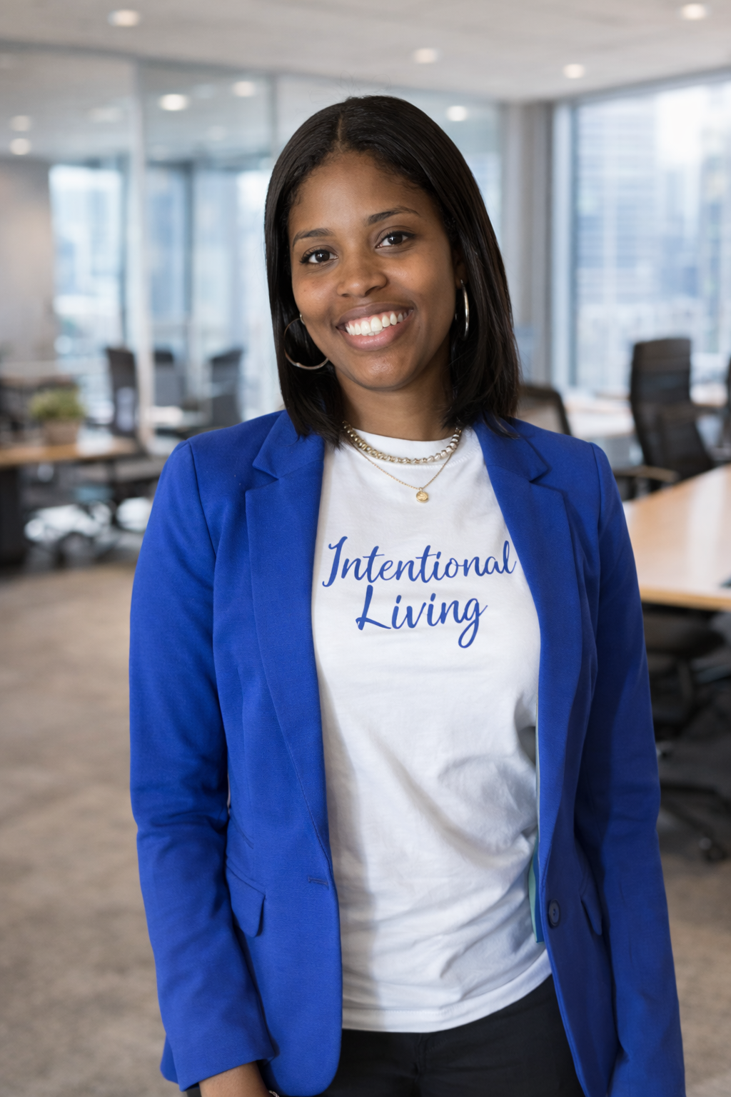 A young woman with shoulder-length black hair wearing a white T-shirt with 'Intentional Living' printed on it, a blue blazer, and jewelry, standing in a modern office with large windows and conference tables.
