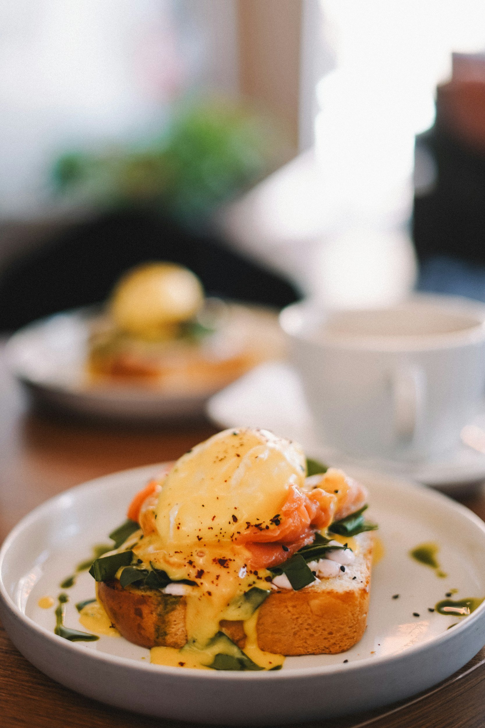 Close-up of eggs benedict with hollandaise sauce, chopped herbs, and black pepper on an English muffin with leafy greens underneath, served on a white plate.