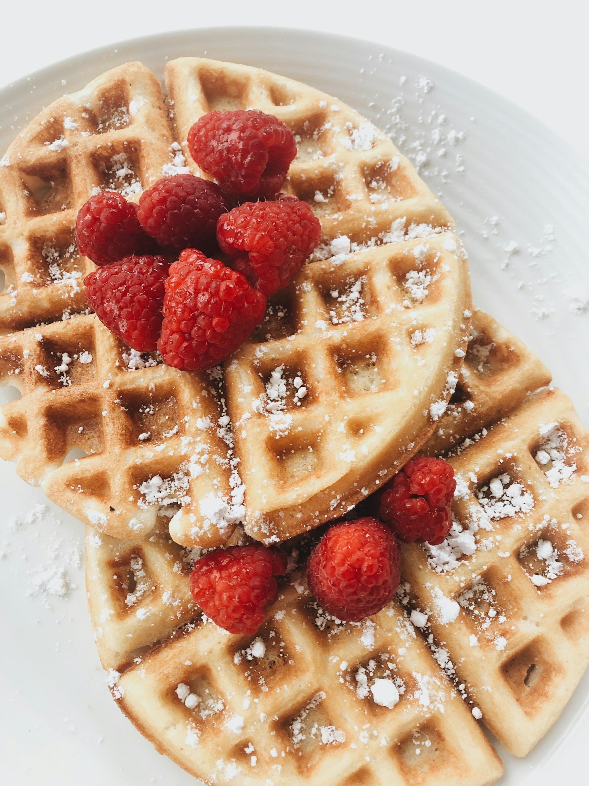 Close-up of two golden-brown waffles topped with fresh red raspberries, powdered sugar, on a white plate.