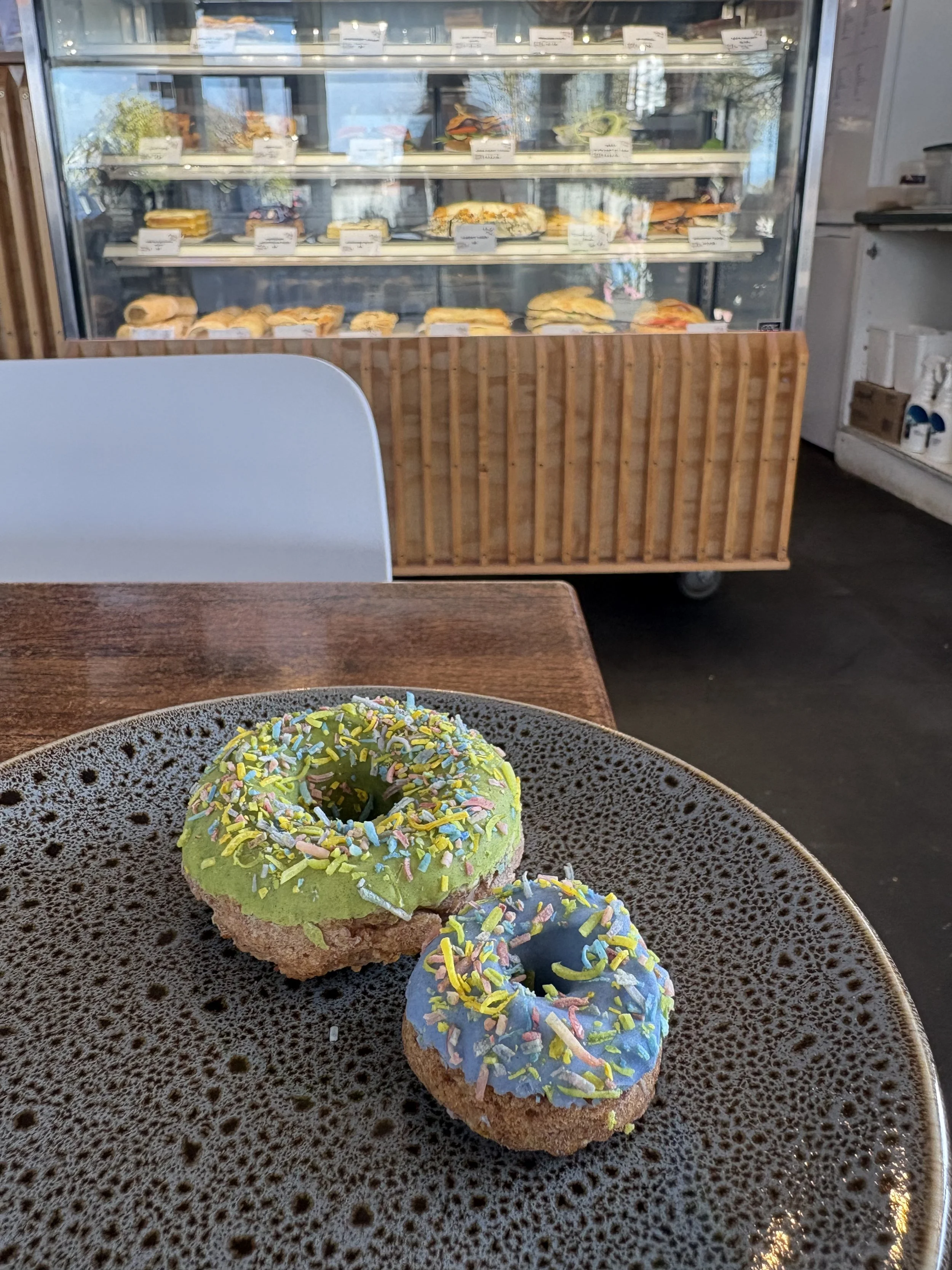 Colorful donuts with sprinkles on a dark, speckled plate in the foreground, with a glass display refrigerator holding various sandwiches and salads in the background.