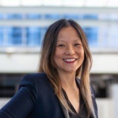 A woman with shoulder-length hair, smiling, wearing a dark blazer, standing outdoors with a blue sky and modern buildings in the background.
