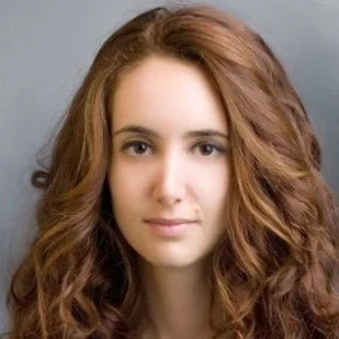 A young woman with long, wavy reddish-brown hair looking at the camera with a neutral expression against a plain background.