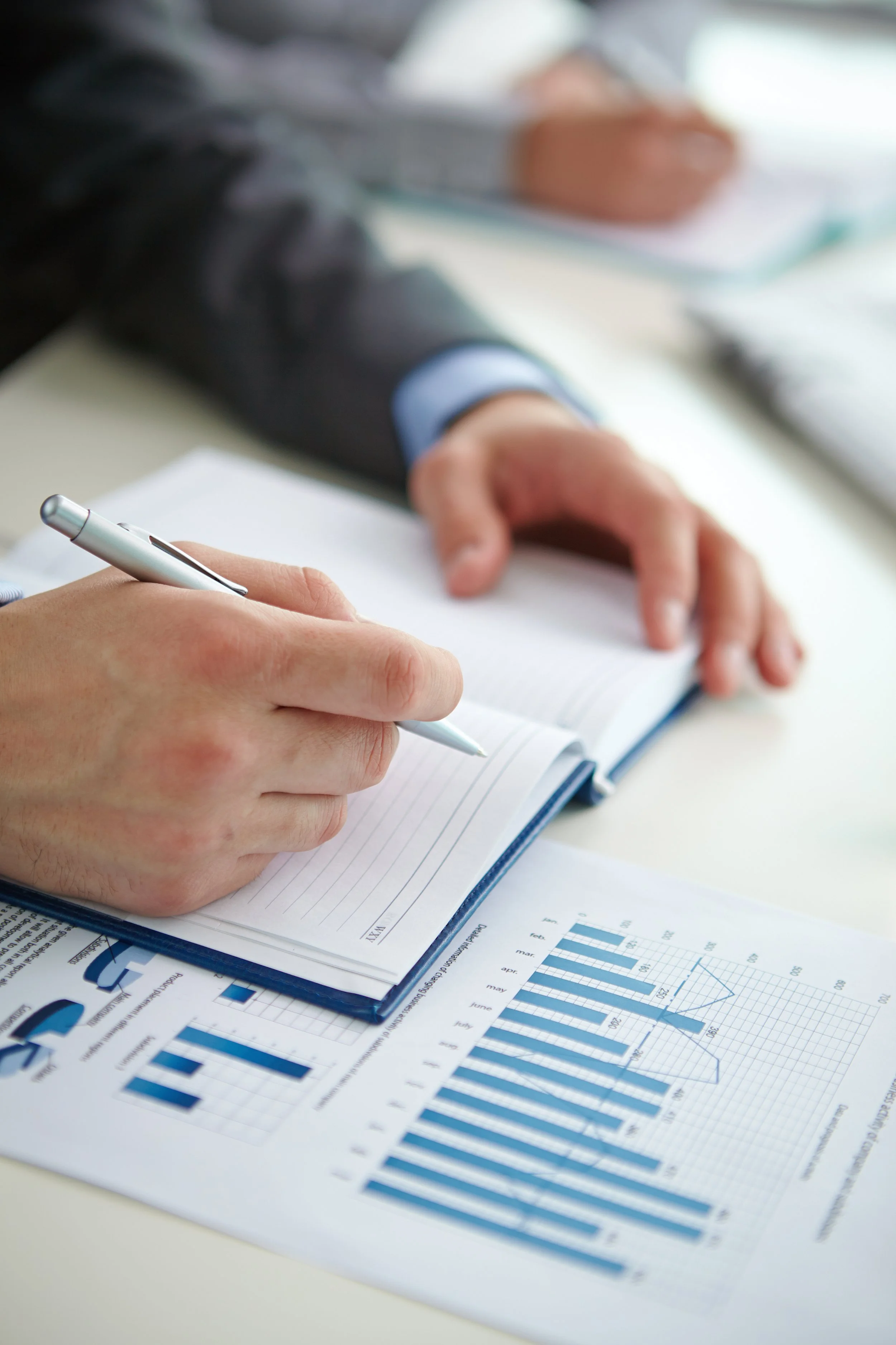 Close-up of a person's hand writing in a notebook, with printed graphs and data sheets on the desk.