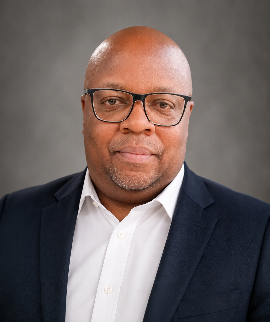 A man wearing glasses, a white shirt, and a navy blazer posing against a plain gray background.