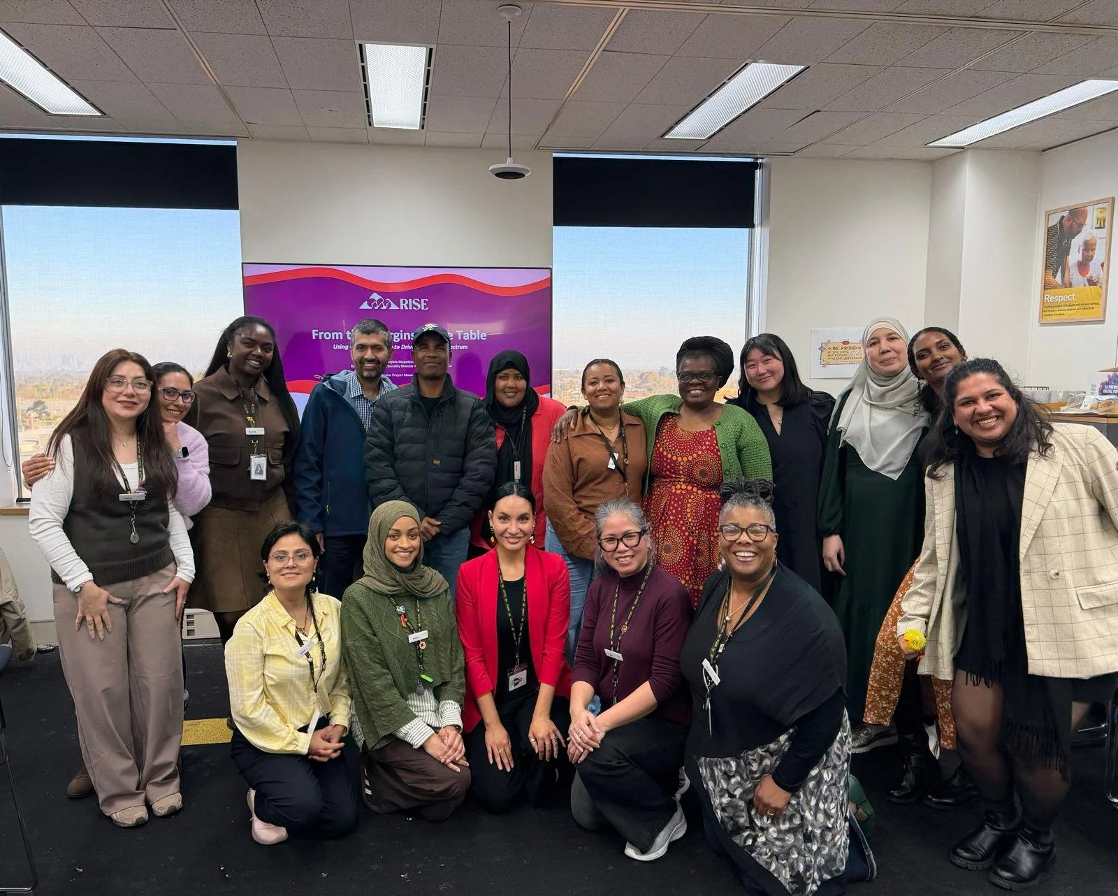 A diverse group of 15 women and men posing together in an indoor office space with windows and a city view in the background. They are smiling and wearing business casual attire, some with name tags. A purple and pink presentation screen in the background displays the word 'RISE' and the phrase 'From the Beginning to the Table.'