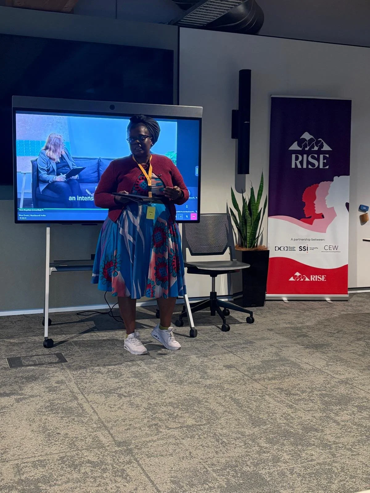 A woman stands in front of a large digital screen and a banner at a conference or event. She is wearing glasses, a colorful patterned dress, a red cardigan, and white sneakers. The woman holds a tablet or notebook and appears to be speaking or preparing to speak. The banner reads "RISE" with a logo and mentions partners, including DCC, SSI, and CEW. There is a potted plant and a chair next to her.