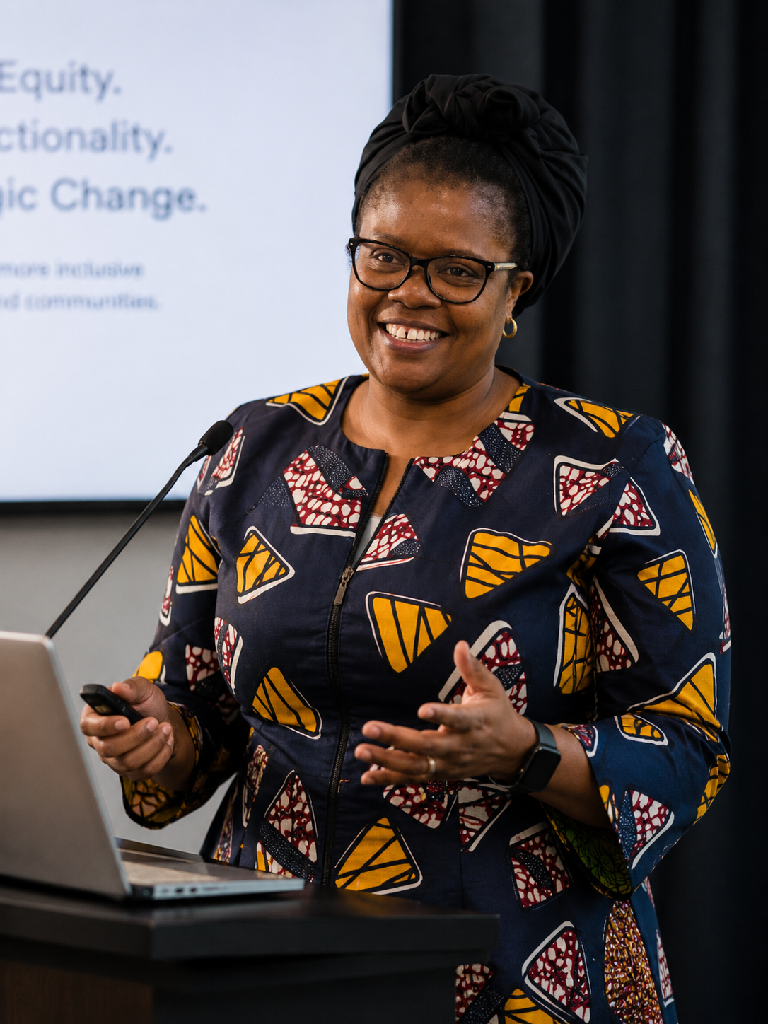 A woman with glasses, a black headwrap, and colorful patterned dress standing at a podium, smiling, holding a remote, with a presentation slide in the background.