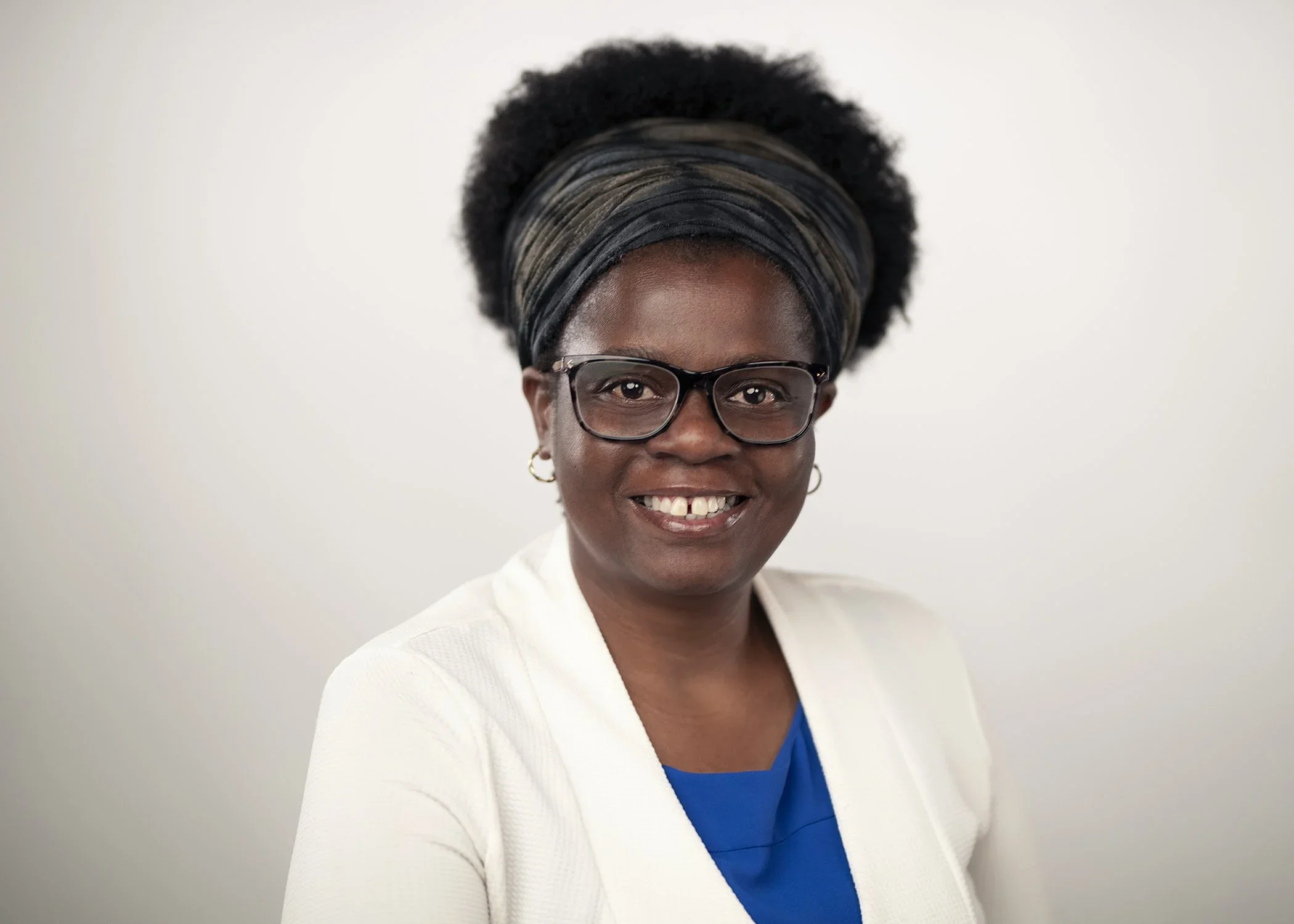 A portrait of a smiling Dr Virginia Mapedzahama . wearing glasses, a headband, white blazer, and a blue top in front of a plain white background, Dr Virginia Mapedzahama .
