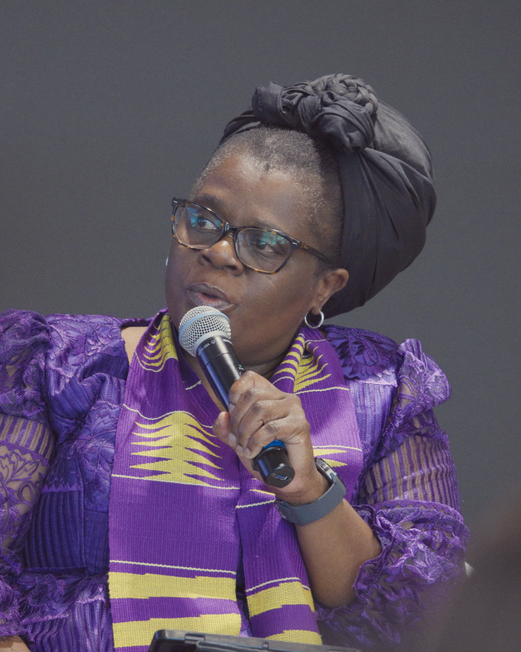 A woman with short hair and glasses speaking into a microphone, wearing a colorful purple and yellow garment with lace details and a black head wrap.