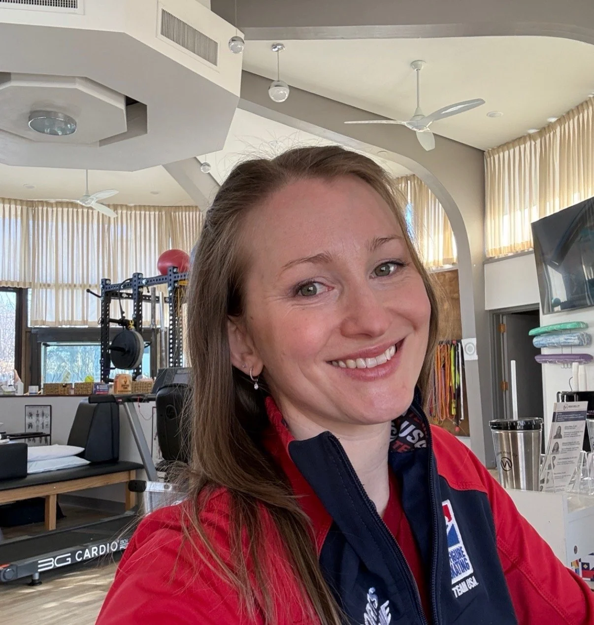 Smiling woman with long brown hair in a gym or fitness room, wearing a red and black sports jacket, with workout equipment and a television in the background.