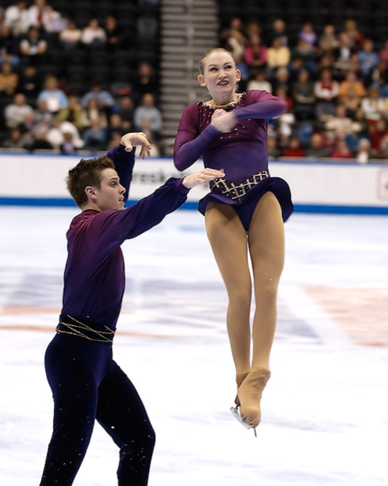 A male and female figure skaters performing a routine on ice, with the female skater mid-air and the male skater supporting her, in front of an audience in an indoor rink.