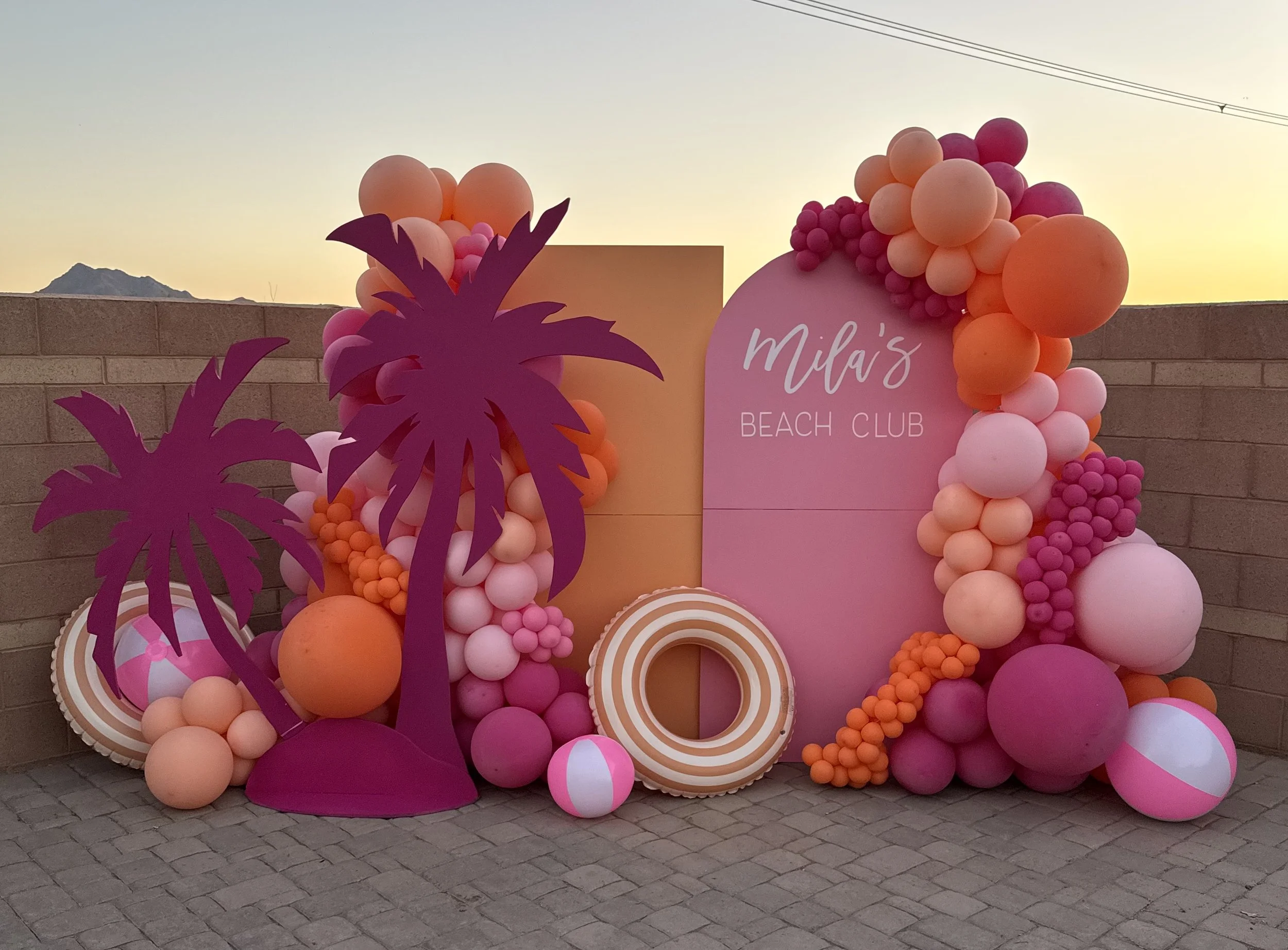 Decorative setup with pink, orange, and purple balloons arranged around a sign that reads "Mila's Beach Club" and purple paper palm trees, set against a low brick wall and a backdrop of a mountain and evening sky.