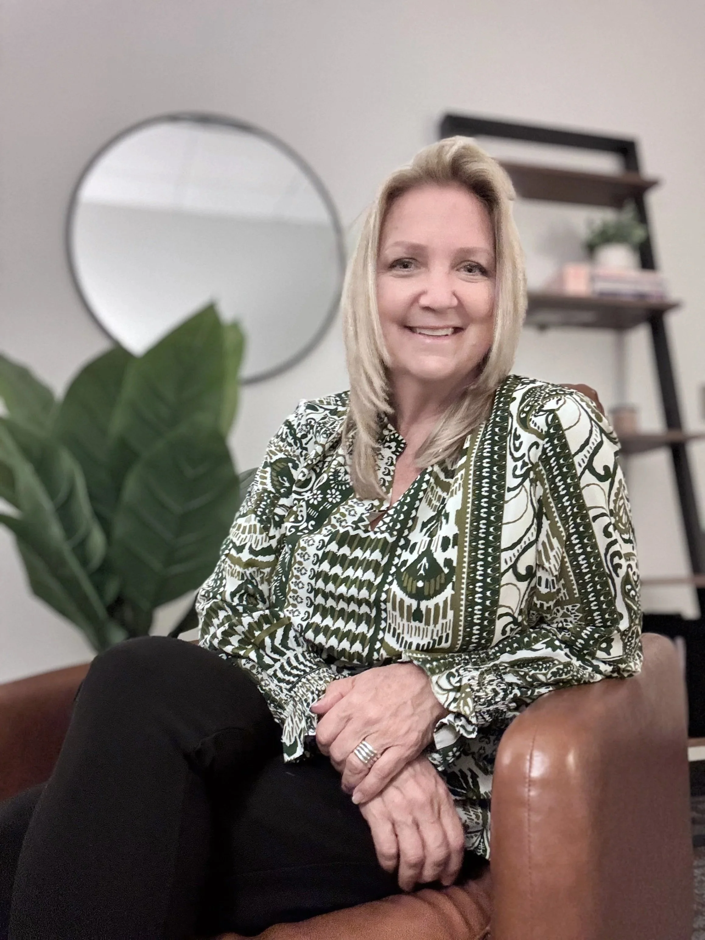 A smiling woman with shoulder-length blonde hair, wearing a patterned blouse, sitting in a room with a mirror, potted plant, and shelves in the background.