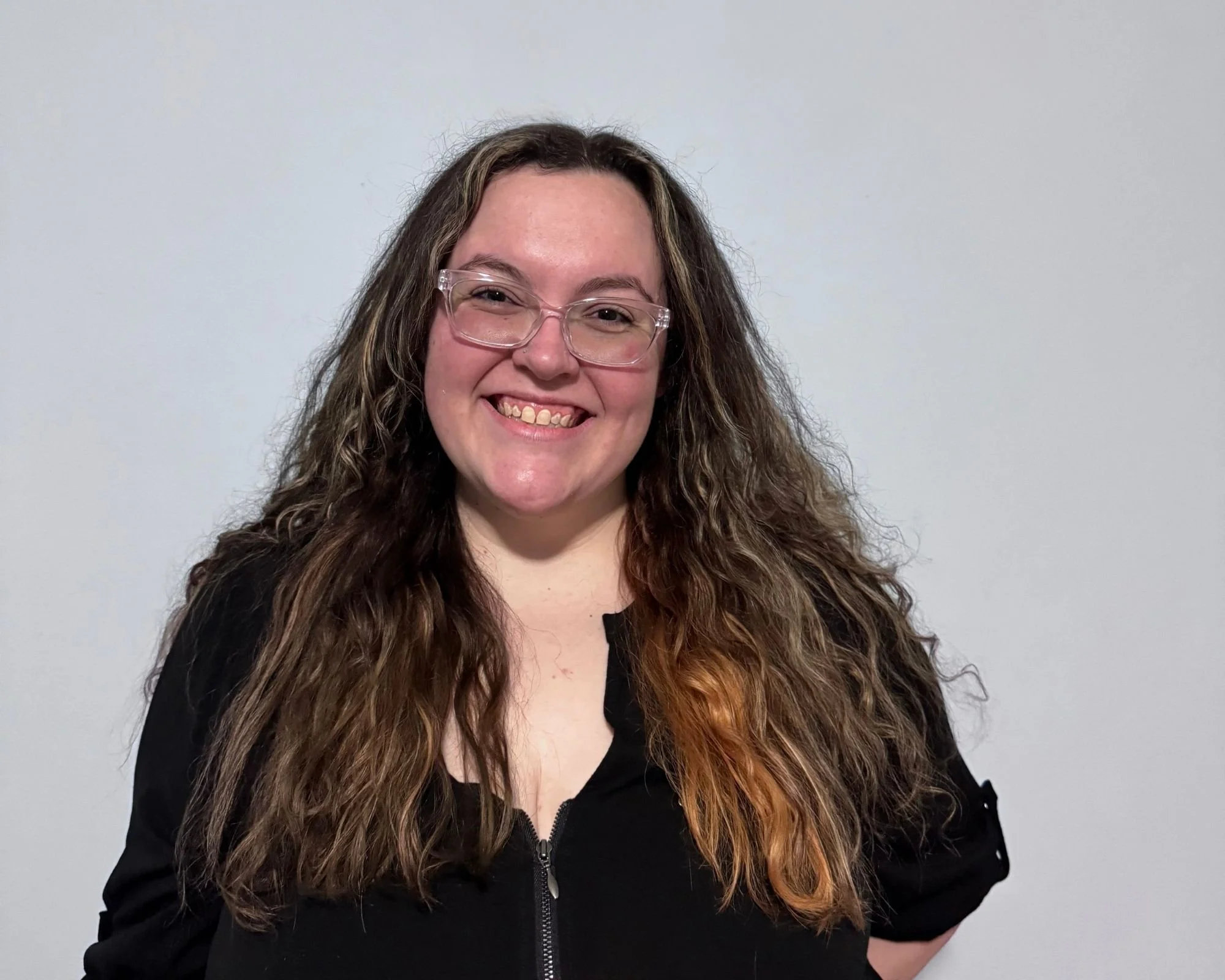 A smiling woman with curly hair wearing glasses and a black zip-up jacket standing against a plain white wall.