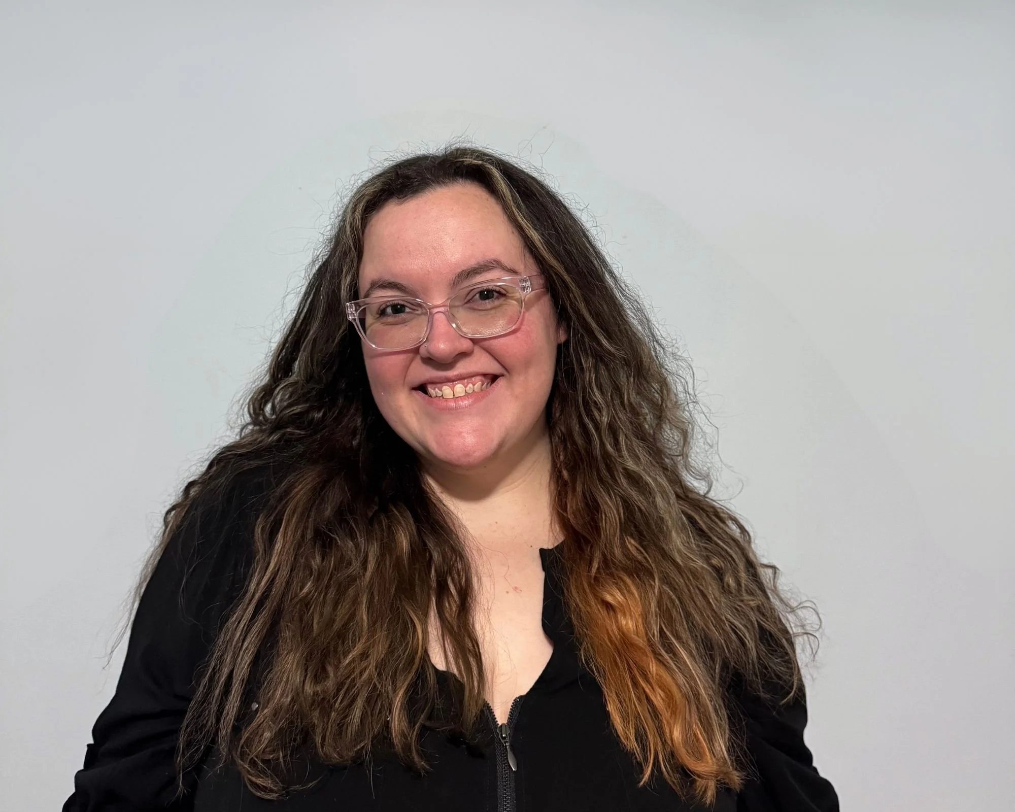 A woman with long curly hair and glasses smiling, wearing a black zip-up hoodie, standing against a plain white background.